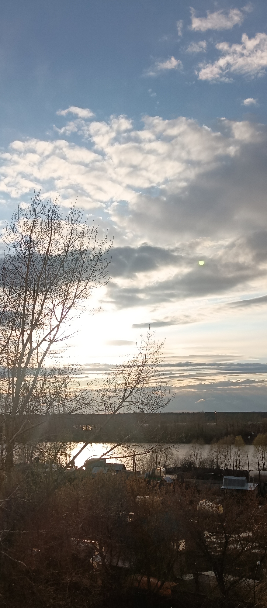 This outdoor landscape image, likely captured from an elevated viewpoint, depicts a serene scene at either sunrise or sunset. The sky dominates the upper portion, featuring a mix of bright white and grey cumulus and stratocumulus clouds set against patches of pale blue. A brilliant, low-lying sun, partially hidden by clouds, emits a strong glare that creates a prominent lens flare, visible as a distinct bright green/yellow circle in the upper right sky.
In the immediate foreground, the stark, leafless branches of a large tree are silhouetted against the bright sky on the left side of the frame. Below this, dense, dark undergrowth and smaller bare trees fill the lower portion, obscuring some details of the ground.
The midground features a wide body of water, likely a river or lake, stretching horizontally across the scene. The intense sunlight creates a powerful, shimmering reflection on its surface. Beyond the water, indistinct structures, possibly sheds or small buildings associated with a rural settlement, are scattered amidst more bare trees and vegetation. The distant horizon is hazy, with a faint line of land or trees visible.
No people or specific activities are visible. The bare trees suggest a season of cold weather, such as late autumn, winter, or early spring. The strong, low angle of the sun clearly indicates either dawn or dusk. There is no visible text in the image.