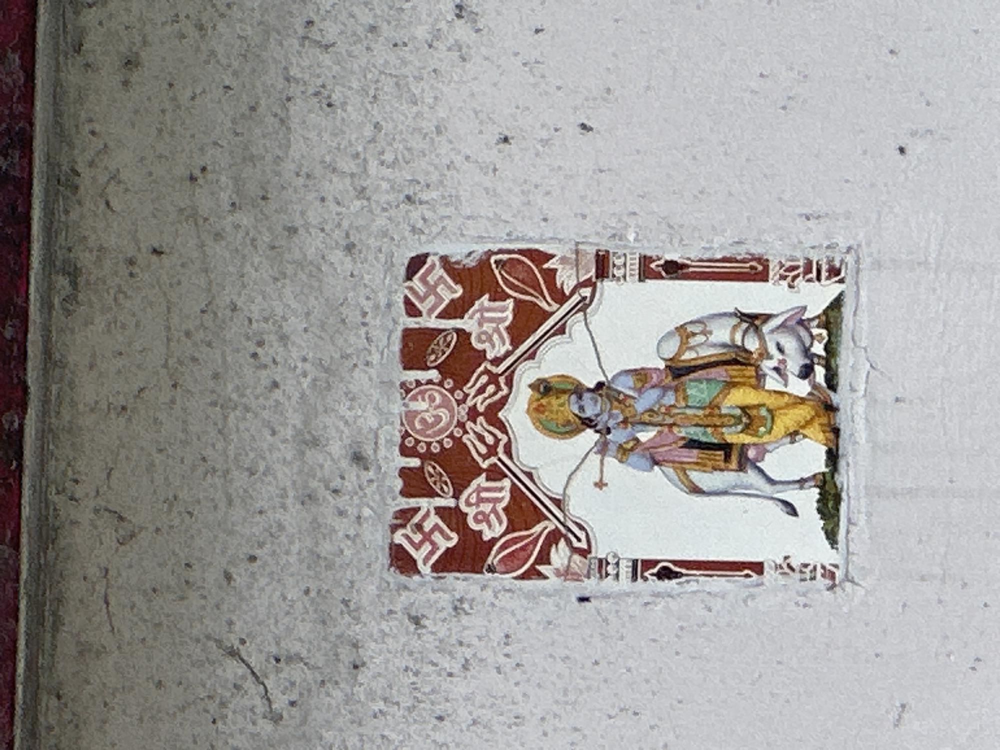A small, tiled image is affixed to a rough, textured concrete wall in Bhagsu, India. The image depicts Lord Krishna playing his flute, riding a white horse. He is adorned in traditional Indian attire, a yellow dhoti, a green and gold vest, and a colorful turban. He is accompanied by a white cow. The artwork is framed by a red border featuring Swastika symbols and the Om symbol. The concrete wall itself shows signs of wear and tear, with cracks and small dark spots scattered across its surface. The top edge of the image is bordered by a dark red stripe.