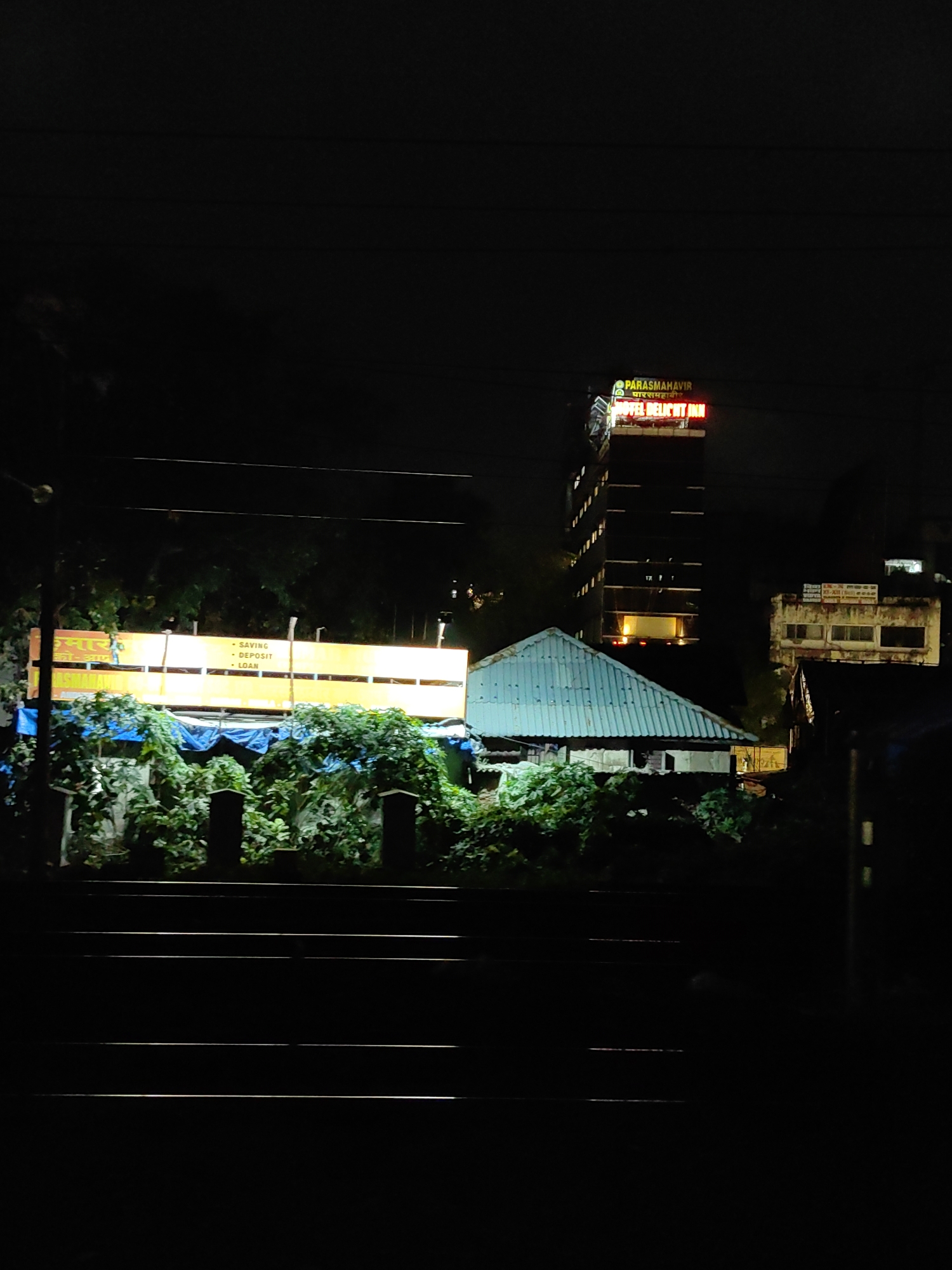 The image depicts a nighttime scene in Mumbai, India, likely near a railway line, given the presence of parallel tracks in the foreground. A prominent neon sign for "HOTEL DELIGHT INN" is visible on a tall building in the background, with the text "PARAMMAHAVIR" also illuminated above it. To the left of the hotel, a brightly lit yellow billboard displays text, including "SAVING", "DEPOSIT", and "LOAN", suggesting it is an advertisement for a financial institution. In the middle ground, a low-rise structure with a blue corrugated metal roof is situated amongst lush green foliage, partially covered by blue tarpaulins. The environment appears to be urban, with evidence of electrical wires strung across the frame, typical of city infrastructure. The overall impression is one of a busy commercial area at night, illuminated by various signs and lights.