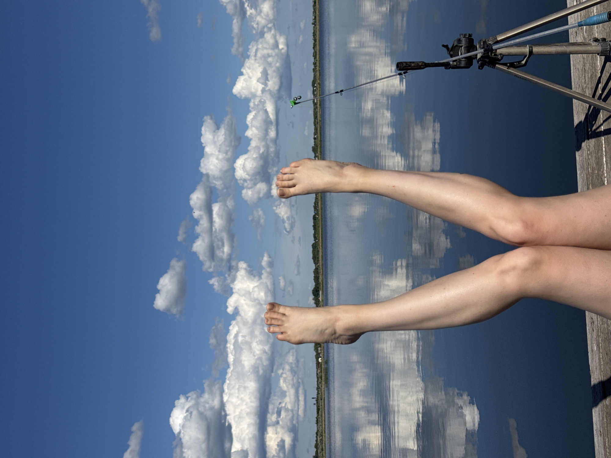 The image depicts a pair of legs and feet extended over a wooden dock. A fishing rod and tripod are positioned on the dock nearby. The background features a body of water reflecting a cloudy sky. A treeline is visible on the distant shore.