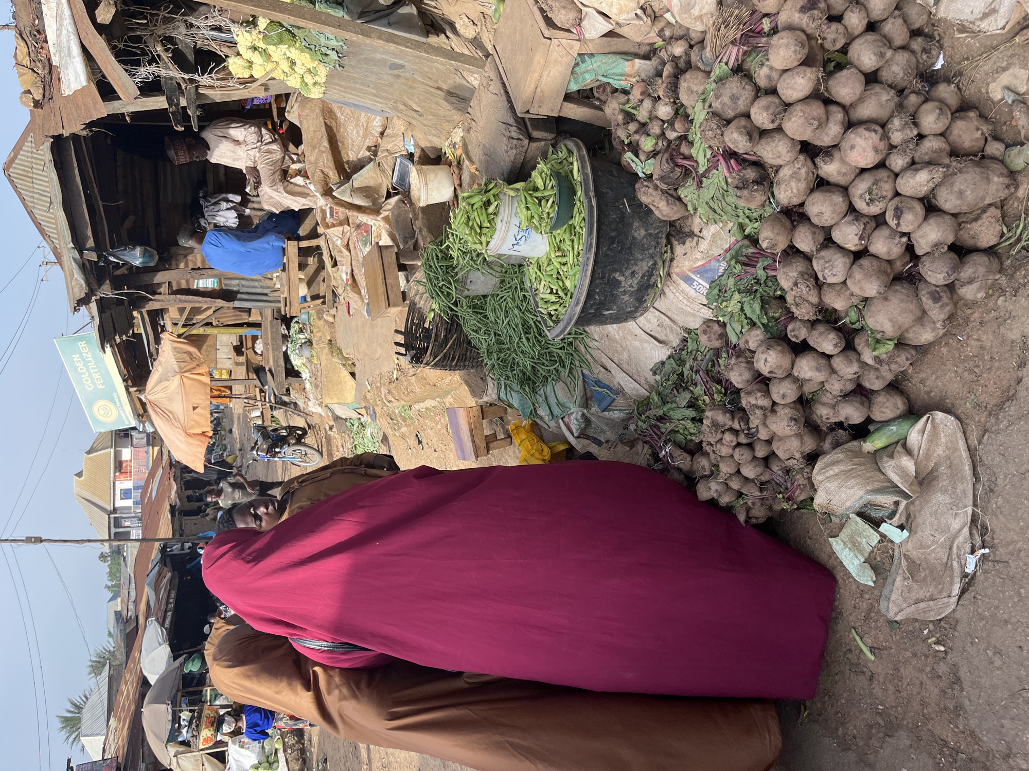 The image depicts a street market scene in Rafin Par, Nigeria. In the foreground, two individuals are partially visible from behind, wearing traditional brown and magenta garments, respectively. To the right, a large pile of beets with their green tops is arranged on the ground, some placed in a burlap sack. Next to the beets, a black plastic basin overflows with green beans, with a smaller green bowl also containing green beans placed on top. A white bucket, also filled with green beans, is situated behind the basin, and a woven basket filled with green beans sits to the left of the bucket. In the background, stalls with corrugated tin roofs are visible, some of which appear to be selling goods. Two men are seated in one of the stalls; one is wearing a blue garment, and the other is in a light-colored, traditional outfit. A motorcycle is parked near the stalls. The ground is dirt and appears uneven. The overall atmosphere suggests a bustling marketplace.