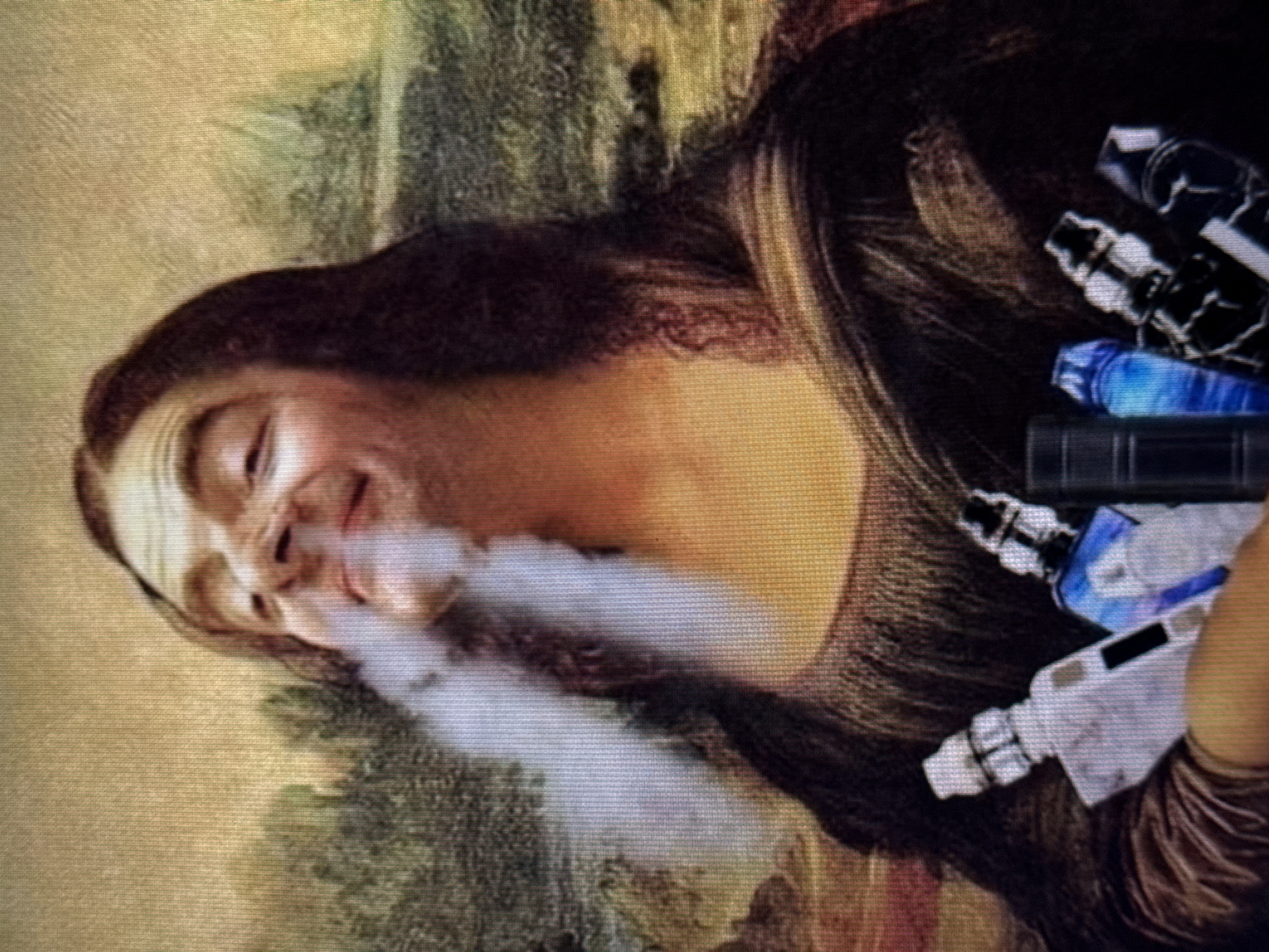 The image shows a digitally altered reproduction of Leonardo da Vinci's "Mona Lisa." The iconic painting is overlaid with modern elements. The subject, Mona Lisa, appears to be exhaling a plume of white vapor, suggestive of vaping. In her right hand, which is not typically visible in the original painting but is added here, she holds several vaping devices. These devices are cylindrical and vary in color, including blue and white, and some have metallic accents. The background remains consistent with the original artwork, depicting a landscape with mountains and a winding path. The lighting suggests an outdoor setting, consistent with the original painting's naturalistic style. The overall impression is a blend of classical art and contemporary culture, creating a humorous or satirical effect. No visible text is present in the image.