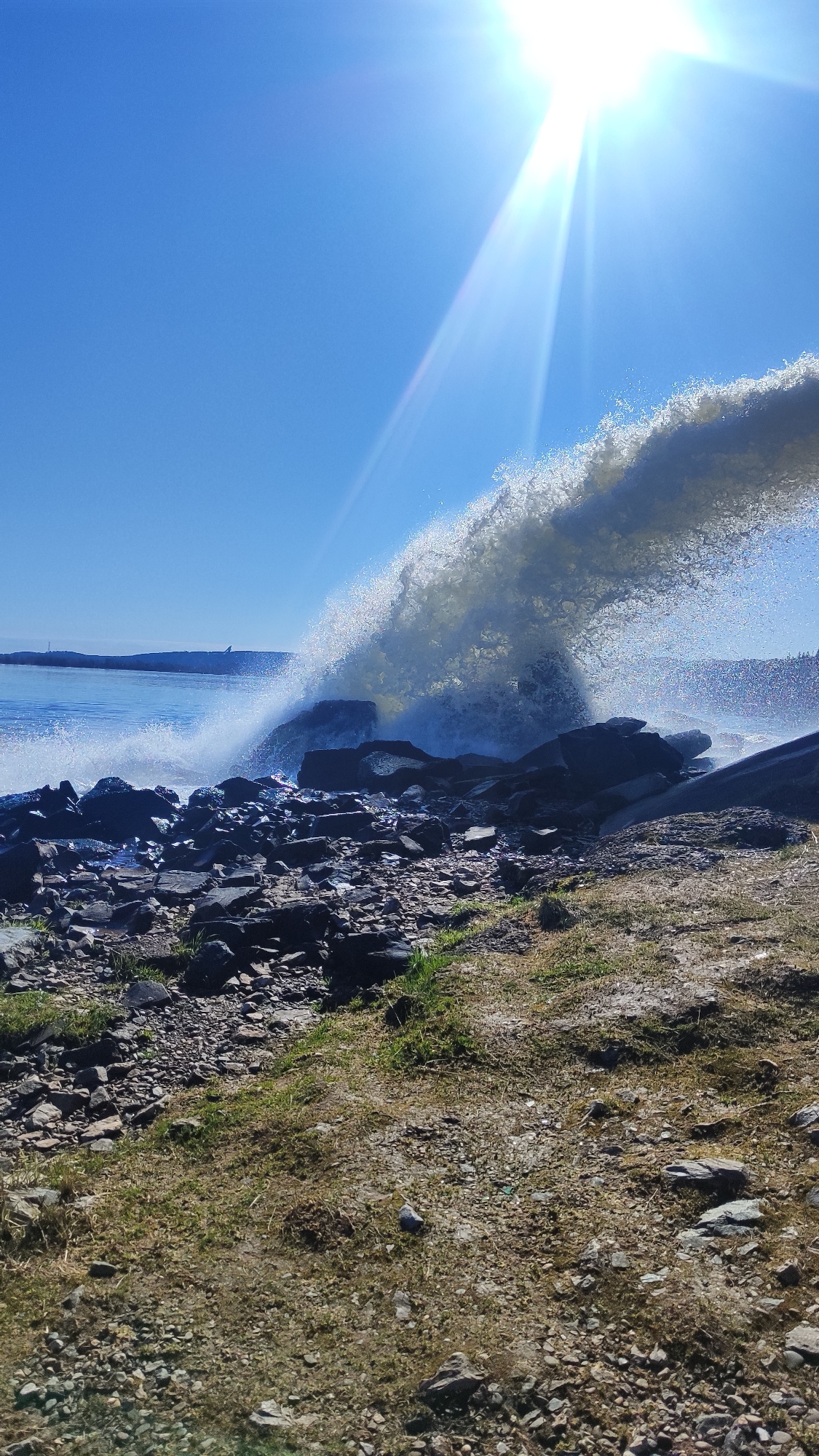 A bright, sunny day illuminates a rocky coastline. A large wave crashes against dark, jagged rocks, creating a spray of water droplets that extends upwards towards the sun. The sky is a clear, light blue. In the distance, a dark blue body of water meets the horizon. On the land, a mix of rocks and grassy patches form the foreground. The photograph does not depict the city of Chernushka, Russia.