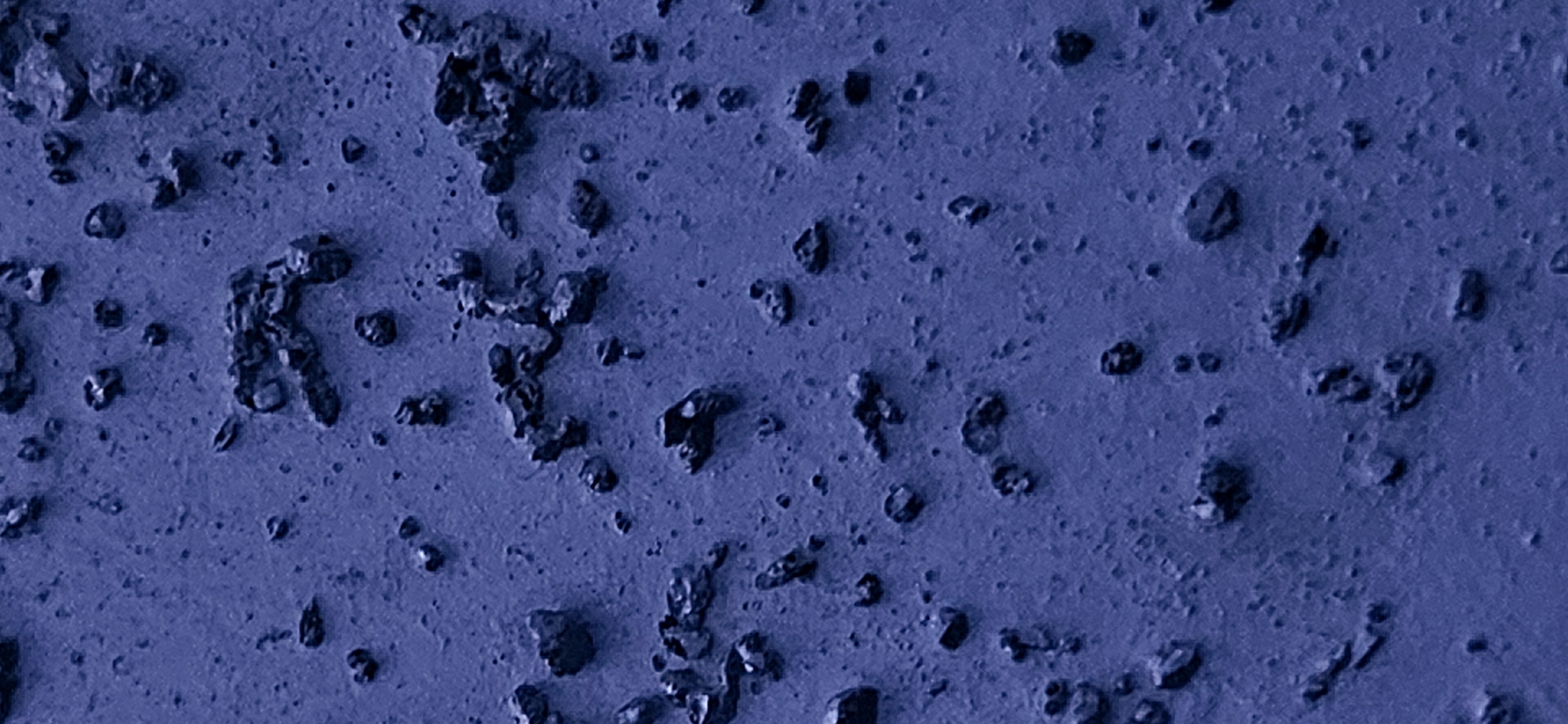 The image displays a close-up, macro view of a textured, blue-toned surface with numerous small, irregular dark-colored particles scattered across it. The surface itself appears to be fine-grained and somewhat porous, with subtle variations in shade and texture creating a mottled effect. The dark particles vary in size and shape, ranging from tiny specks to larger, irregularly clumped fragments. Some appear embedded in the surface, while others sit on top. The overall impression is that of a granular or rocky material. The scene is devoid of any human presence or discernible structures, suggesting an abstract or naturalistic subject. The location specified, Momin Prohod, Bulgaria, does not provide specific visual context for this abstract image, as the image itself does not contain any identifiable landmarks or features that would confirm its origin in that particular place.
