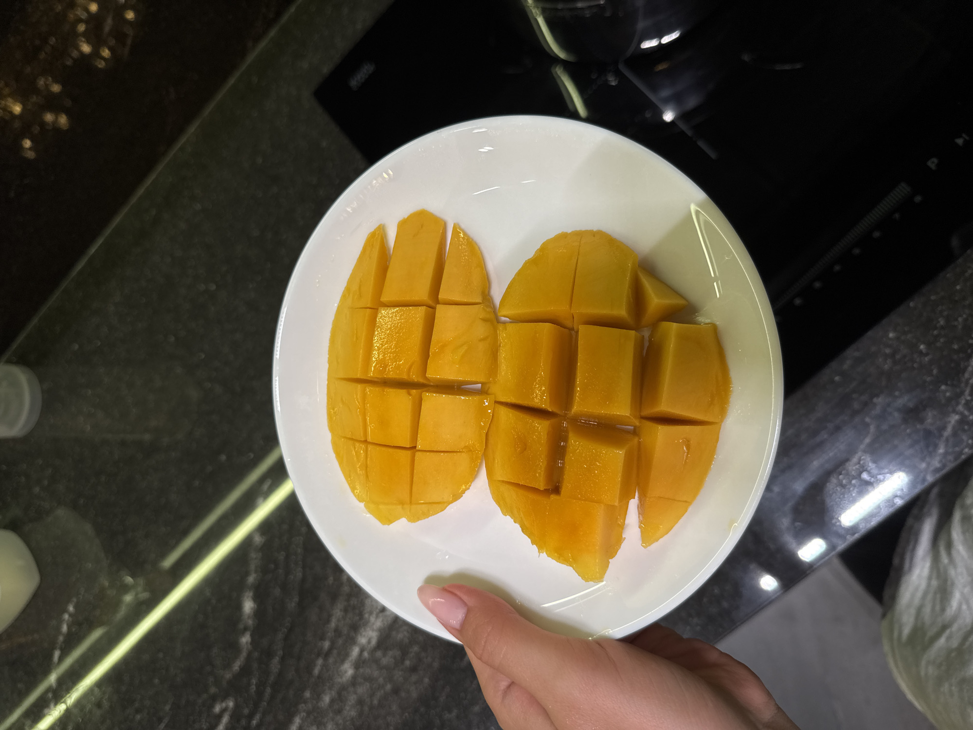 A top-down shot shows a white plate holding two halves of a mango, each scored and cut into cubes, sitting on a dark, reflective countertop. A hand holds the plate. The countertop has light-colored streaks and speckles. In the background, a black induction cooktop and other kitchen items are visible. The location is in a kitchen in an unknown city.