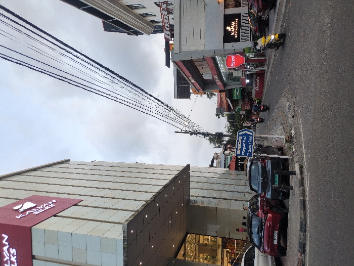 The image displays an urban street scene on Dhanwantari Road in Ernakulam, India, under an overcast sky. On the left, a multi-story commercial building, identified by prominent red signage as "KALYAN SILKS," features a light-colored tiled facade and a large glass-fronted ground floor entrance. A white SUV, a red sedan (Hyundai i20 N Line with partial license plate "KL 07"), and a dark-colored hatchback are parked along the left curb. Figures are vaguely visible within the glass entrance of Kalyan Silks.
In the center foreground, a blue rectangular street sign with a white arrow points right, indicating "DHANWANTARI ROAD" in both English and Malayalam script. A utility pole with numerous overhead power lines spans across the upper center of the frame. The asphalt road, which appears slightly damp, curves to the right.
The right side of the road is lined with multiple commercial buildings. Visible establishments include a business with a red vertical sign labeled "MAYURI" and another sign for "KANAI RESTAURANT." A yellow scooter is moving towards the viewer on the right lane, while two other motorcycles or scooters are further down the road, heading away from the viewer. Several additional cars are parked along the right curb. The overall environment is a bustling commercial area with moderate traffic.