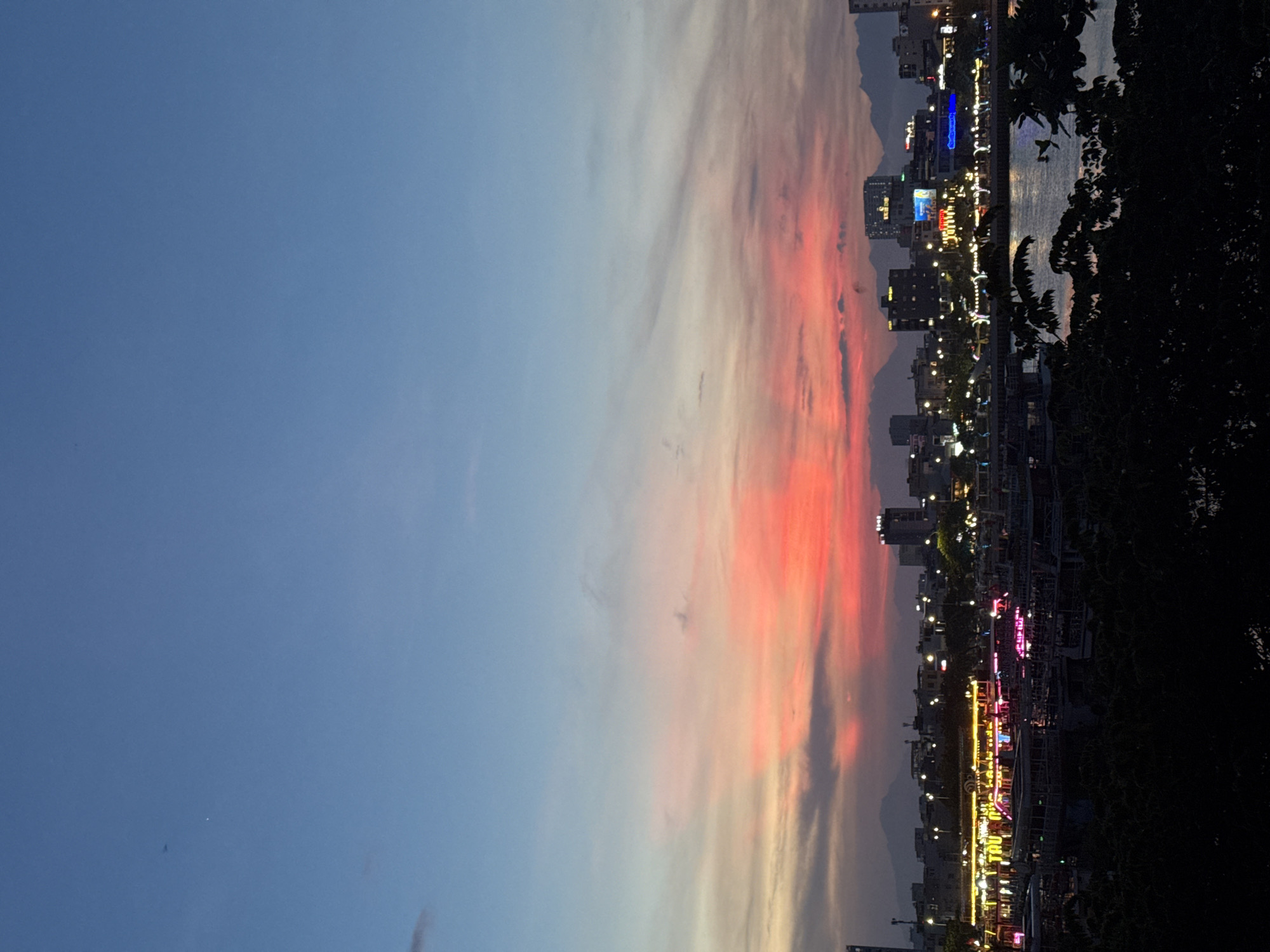 The image captures a vibrant sunset over the city of Đà Nẵng, Vietnam. The sky transitions from a deep blue at the top to fiery orange and pink hues near the horizon, with streaks of clouds painting the sky. In the foreground, the Han River flows, reflecting the colorful sky and the lights of the city. Several boats are docked along the riverbank, some adorned with bright, colorful lights, suggesting a lively evening scene. The cityscape is illuminated by numerous lights, with buildings and silhouetted mountains forming the backdrop. The overall impression is one of a bustling city at dusk, bathed in the warm glow of a dramatic sunset.