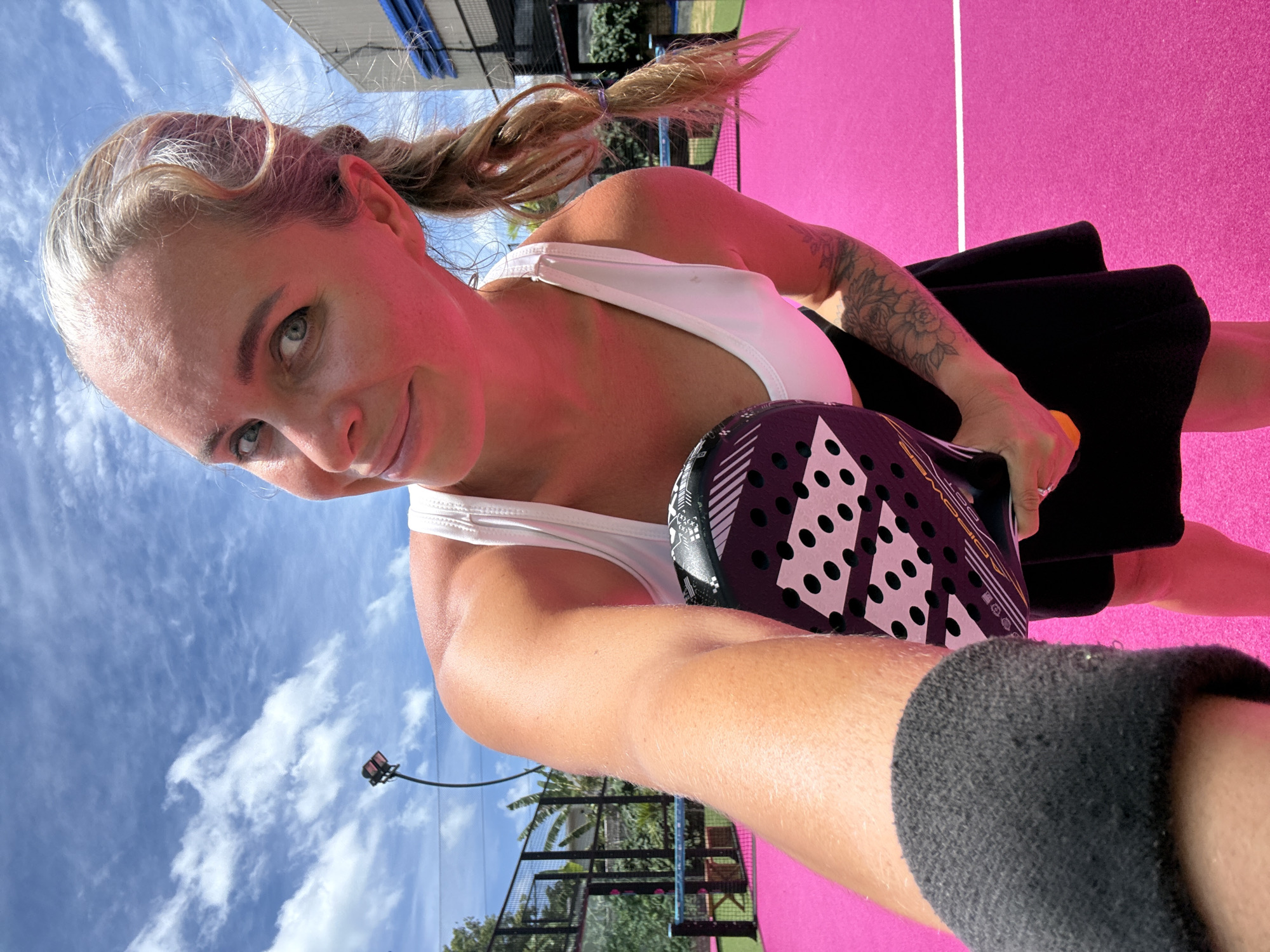 In Ko Samui District, Thailand, on a brightly lit outdoor padel court with a vibrant pink surface and white boundary lines, a woman with blonde hair pulled into a braid stands holding a black padel racket with a white dot pattern. She is wearing a white tank top and a black skirt. Her left arm is extended towards the camera, with a gray sock visible on her left foot. The background features a net, fencing, trees, and a bright blue sky with scattered white clouds. A spotlight is visible in the distance. The woman is looking at the camera with a slight smile.