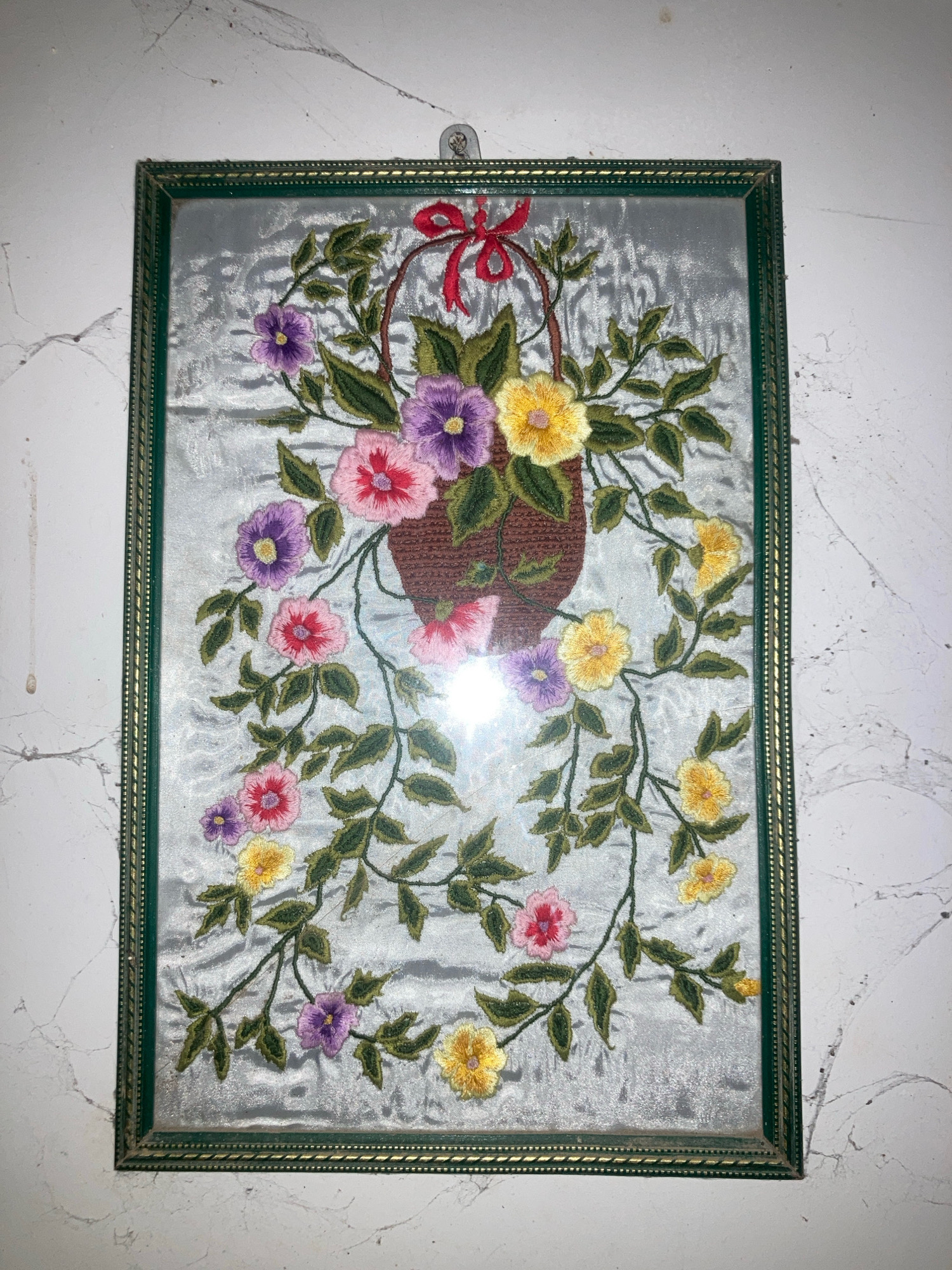 An embroidered floral basket hangs on a textured white wall. The artwork is framed by a dark green and gold border. The basket, depicted in brown embroidery, overflows with colorful flowers in shades of pink, purple, and yellow, intertwined with green leaves and vines that trail down the sides of the frame. A red ribbon is tied in a bow at the top of the basket. The wall exhibits a mottled texture with subtle grey veining, and a thin spiderweb is visible in the upper left corner. The overall impression is of a delicate, handmade piece of art. This image was captured in Daladagama, Sri Lanka.