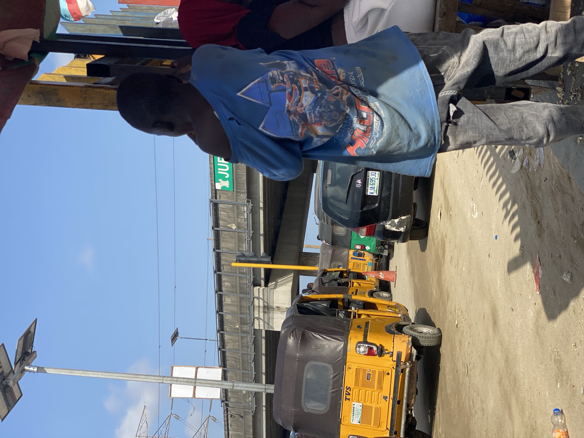 The image shows a street scene in Lagos, Nigeria. A person wearing a blue t-shirt with a graphic design and gray pants is standing to the side. In the street are several yellow three-wheeled vehicles called "keke", typical in Lagos. A black car is behind them. An overpass is visible in the background. The license plate number of the black car is KJA-695-JO. A sign saying "JUP" is visible. A street light and power lines are on the left side of the image.