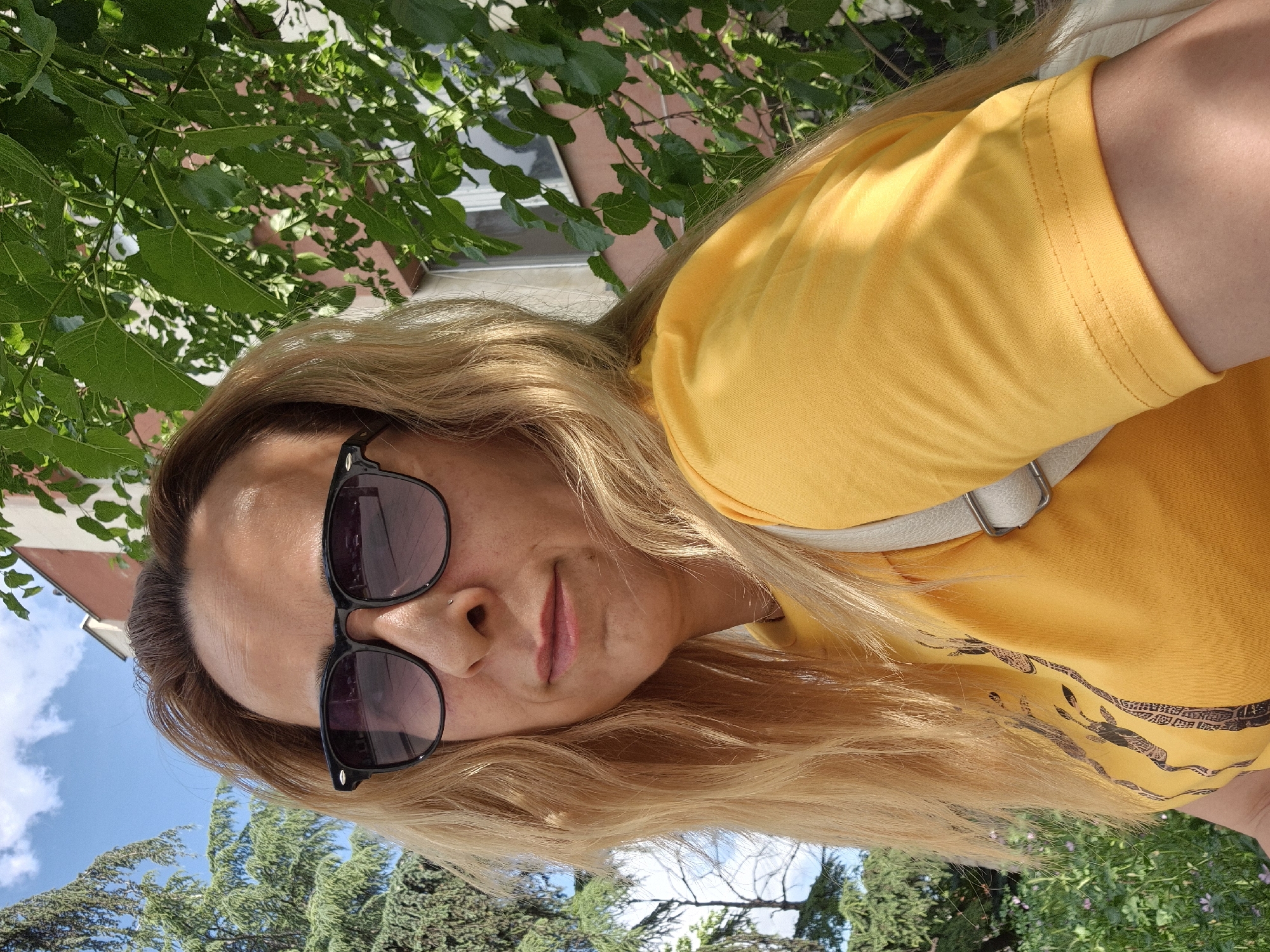 The image is a rotated close-up portrait of a person outdoors, captured on a bright, sunny day. The original orientation appears to be a vertical selfie or shot from a low angle, showing the subject from the chest up.
The subject is a light-skinned individual with long, flowing blonde hair. They are wearing dark, rectangular-framed sunglasses that cover their eyes. A small nose piercing is visible on their right nostril. Their lips are painted a light pink and are slightly parted. The person is wearing a bright, short-sleeved yellow t-shirt. On the lower right side of the t-shirt, there is a distinct dark pattern composed of what appear to be stylized figures or symbols. A light-colored strap, possibly from a bag, is visible across their chest/shoulder.
The background is a mix of natural and urban elements. Lush green foliage, likely from trees and bushes, is prominent, appearing both above and below the subject's head. Through the leaves in the upper background, a section of a reddish-brown building with windows is visible. The lower background shows a clear blue sky with a few scattered white clouds. The lighting is strong and natural, suggesting it is daytime.
No discernible text is visible in the image.