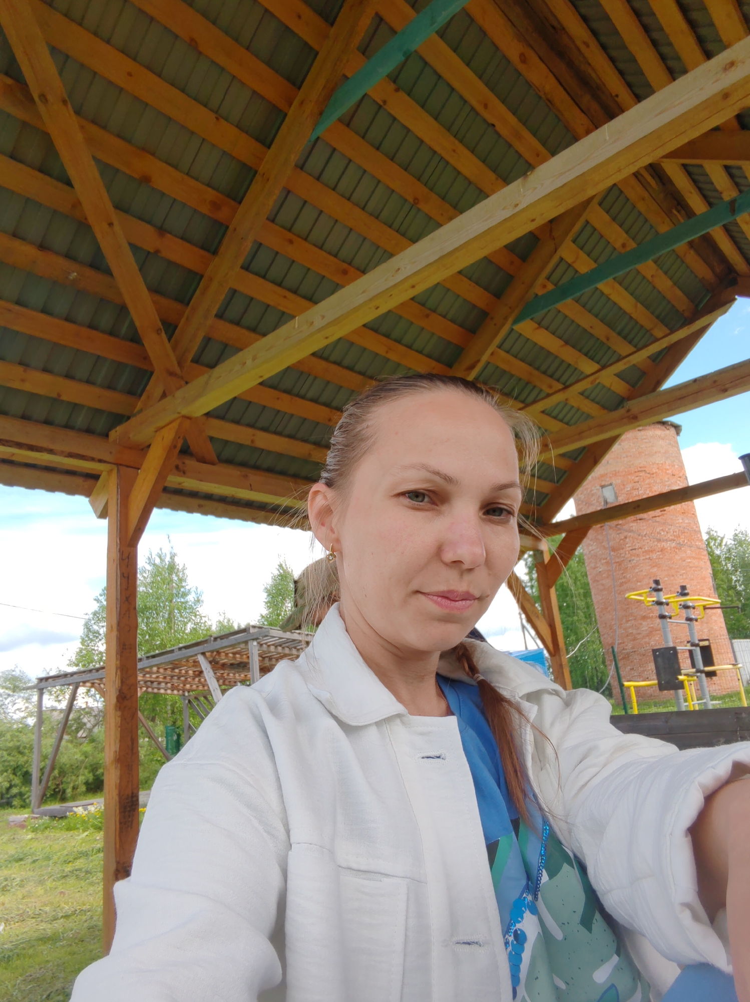 A selfie of a woman with her hair pulled back and wearing a white linen jacket, standing under a wooden structure with a green metal roof. The location is in Чухлэм, Russia. Behind her is a brick tower with exercise equipment in front of it, and other wooden structures are visible in the background. The sky is partly cloudy.