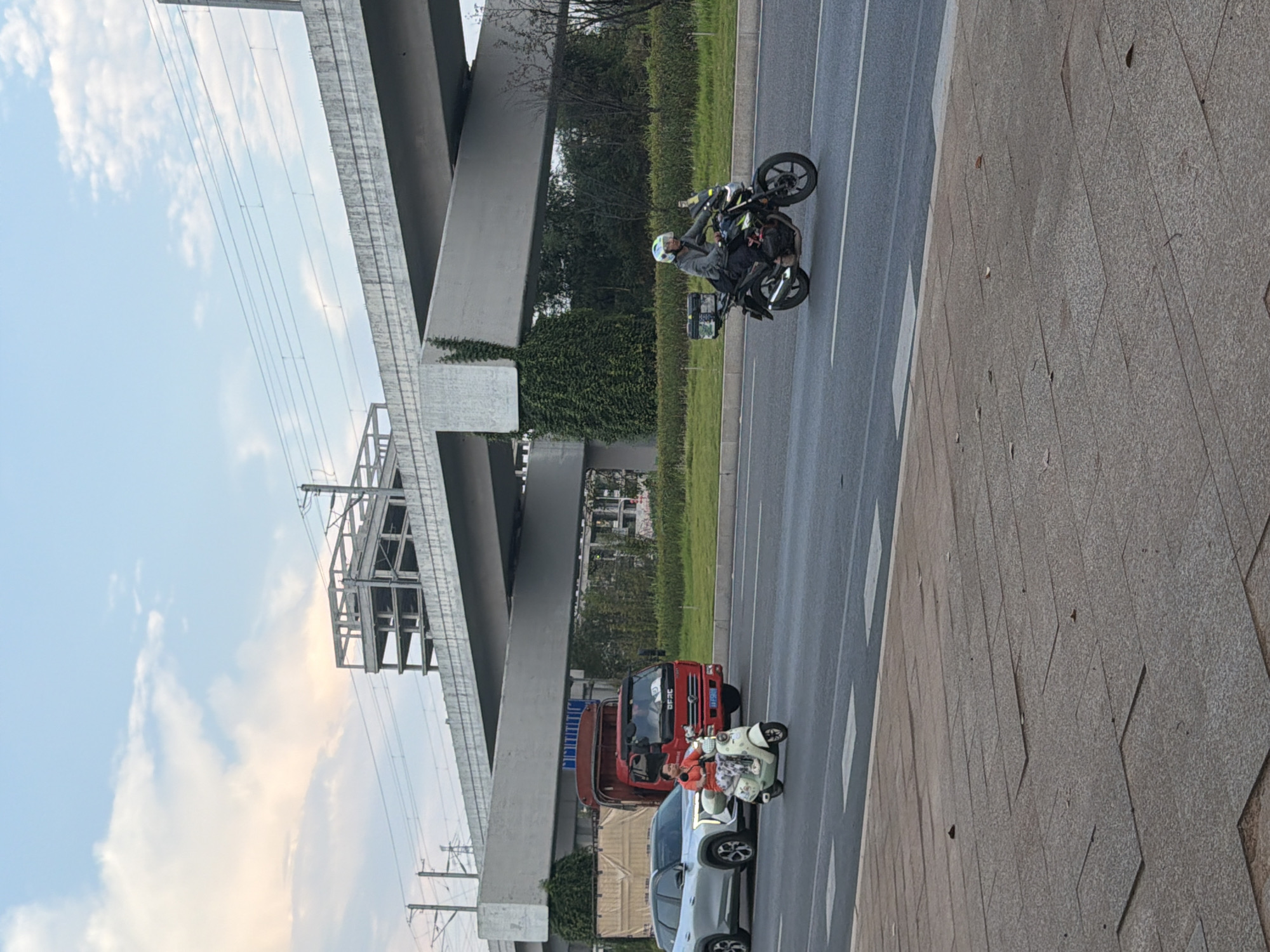 The image shows a multi-lane road with traffic under an elevated highway structure. In the foreground, a person rides a light-colored scooter, wearing a red top and patterned pants. Behind the scooter, a red truck is visible, with its license plate obscured. To the left of the scooter, a silver SUV is parked or moving slowly. Further down the road, a person in a grey jacket rides a black motorcycle with a storage box on the back. The elevated highway is a concrete structure with visible rebar, and electrical wires run above it. The sky is partly cloudy with hints of pink and orange, suggesting either sunrise or sunset. The road surface is asphalt, marked with white lines. The sidewalk on the left is paved with large granite-like tiles. The environment appears to be an urban or suburban area in Jiangjin, China, characterized by modern infrastructure.