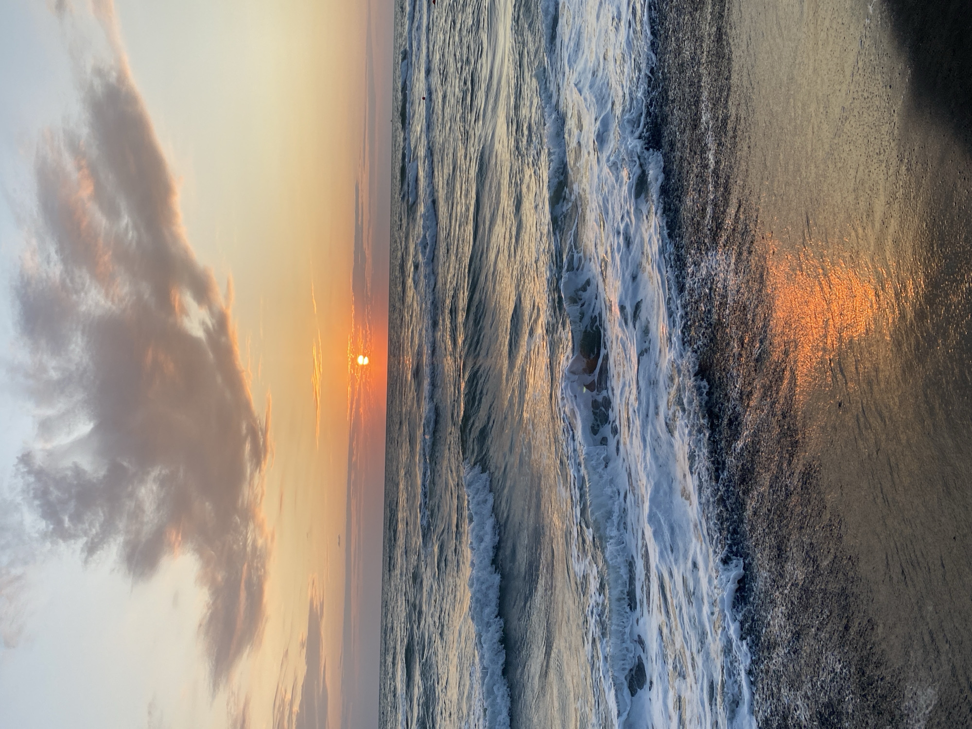 A vertical, eye-level, full shot captures a beach scene in Ureki, Georgia, during sunset. The sun, a bright orange orb, is partially obscured by clouds and hovers just above the horizon line in the upper center of the frame. The sky transitions from a pale blue at the top to shades of orange and pink as it nears the horizon, illuminated by the setting sun. Wispy, pink-tinged clouds stretch across the upper portion of the sky.
The sea fills the majority of the image, with gentle waves rolling towards the shore. The water is a deep blue-green, reflecting the warm hues of the sunset. White foam crests the waves, particularly in the foreground where they are breaking on the dark, pebbly beach. The sand itself appears damp and dark, glistening with the reflection of the sunset.
There are no distinct people or identifiable objects in the water or on the beach, other than the natural elements of the sea, sand, and sky. The overall atmosphere is serene and tranquil, emphasizing the natural beauty of the coastal landscape at dusk.