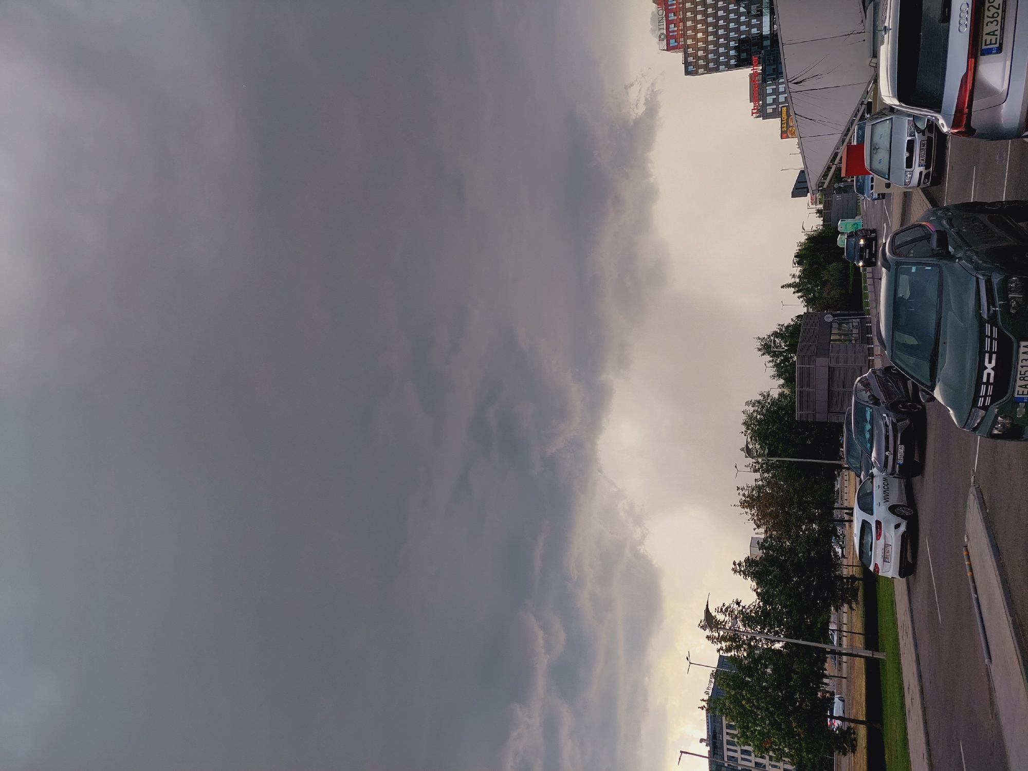This is a photograph taken from a street-level perspective in the city of Sofia, Bulgaria, possibly in the business district. The sky is heavily overcast with dark, dramatic storm clouds dominating the upper half of the frame. A faint rain shower appears to be occurring in the distance.
In the foreground, a paved road is visible with several cars parked and driving. A dark green Dacia Duster is parked in the lower right corner, facing left. To its right and slightly behind it, a silver Audi SUV is parked, facing right, with its license plate "EA 3629" partially visible. Further back, a silver Skoda is parked. To the left of the Audi, a dark-colored SUV, possibly a Renault Austral, is parked. A white Kia Ceed with "VIVACOM" branding is parked in the middle ground, facing left. A darker SUV is parked next to it.
On the left side of the image, a modern office building with many windows is situated behind a row of green trees. Streetlights are also visible along the road. On the right side, a tall, modern building with glass windows and visible signage, including "TINO" and "THERMO FISHER SCIENTIFIC", stands prominently. A white, curved structure, possibly part of a building or a large vehicle, extends across the mid-right of the frame. The overall scene suggests a typical urban environment on a day with impending or ongoing inclement weather.