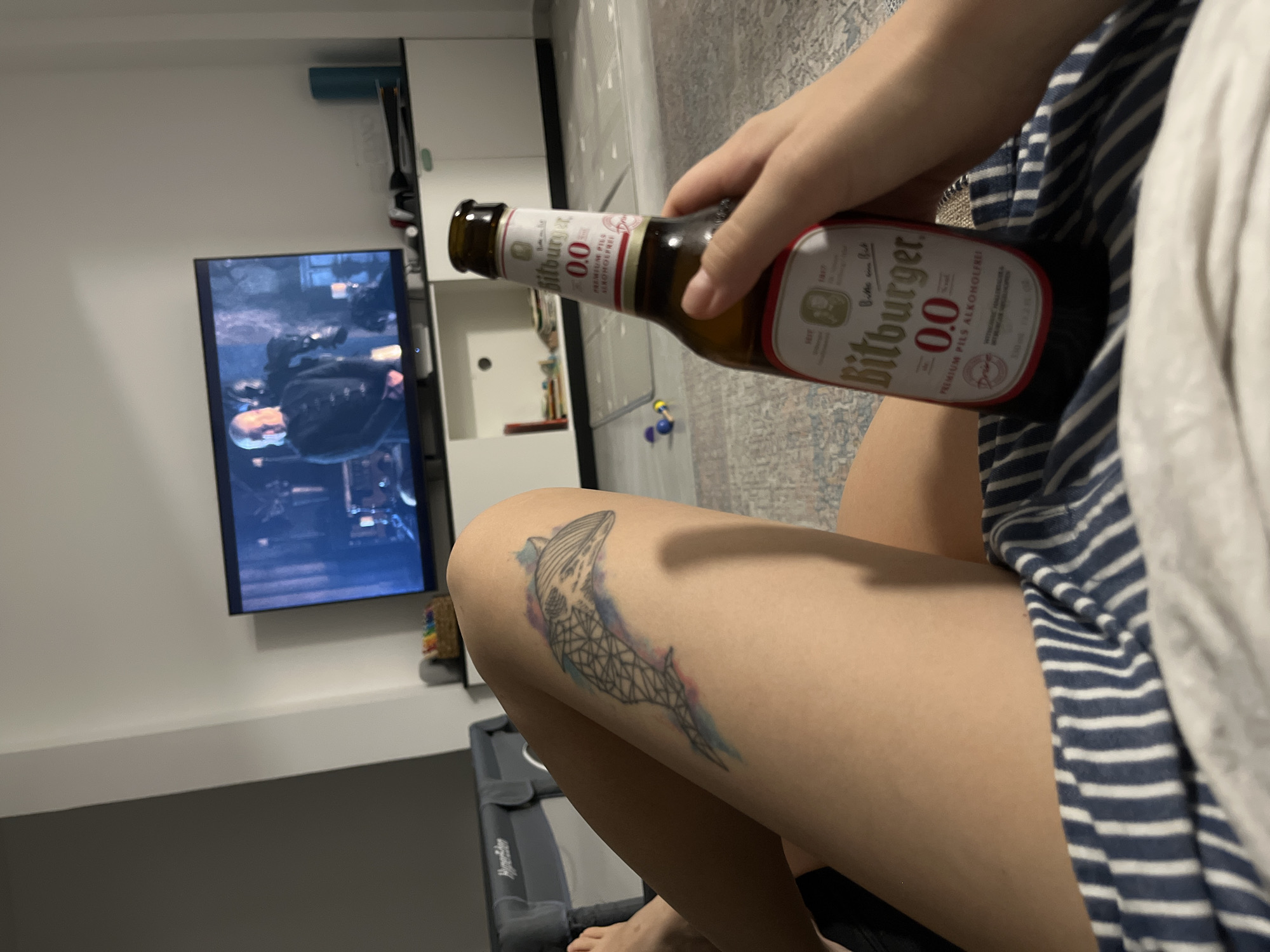 The image depicts an indoor scene, likely a living room, where a person is relaxing. A television screen displays a scene with a man in a dark outfit. In the foreground, a bottle of Bitburger 0.0% premium pils is held by a person's hand. The person's legs are visible, showing a tattoo of a geometric whale with watercolor accents on their left thigh. The individual is wearing striped pajamas. In the background, a gray baby crib and a white cabinet are visible. The overall setting suggests a casual, home environment. The image does not contain any specific elements that would indicate a location in Limassol, Cyprus.