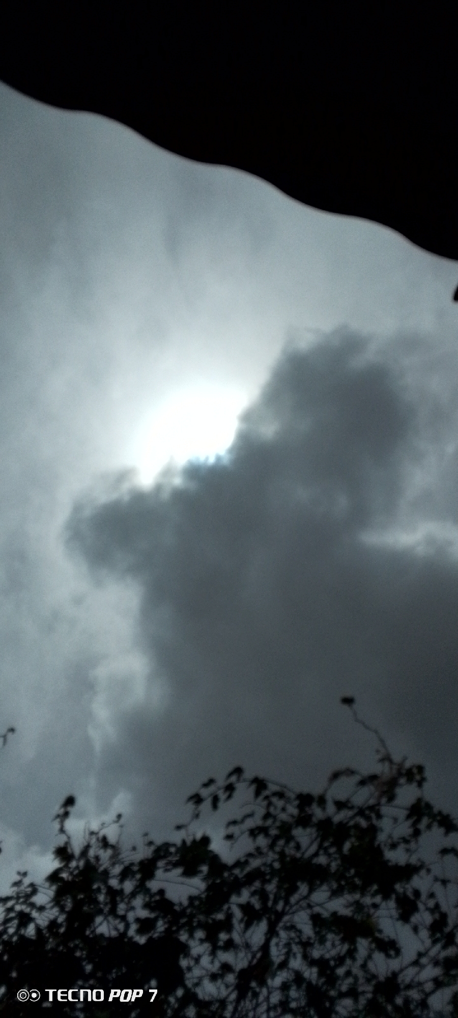 The image shows a view of the sky in Altagracia, Venezuela, as seen from below. The sky is filled with dark, heavy clouds. The sun is visible, partially obscured by the clouds, casting a diffused light through them. In the lower portion of the frame, there's a silhouetted tree. The top of the frame is dominated by a dark, unidentifiable object, possibly a roof or overhang. The image was taken using a Tecno Pop 7.