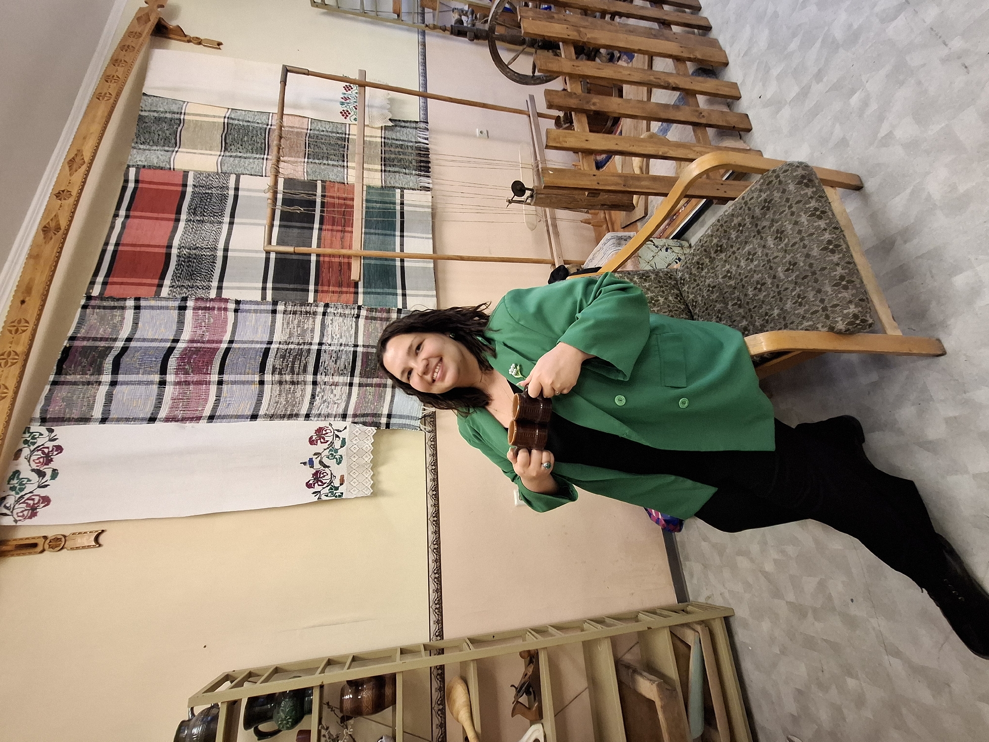 A woman with dark hair, wearing a vibrant green jacket over black clothing, smiles warmly at the camera while holding two small brown ceramic cups in her hands. She is positioned slightly to the right of the frame, appearing cheerful.
Behind her, the scene is rich with elements of traditional craftsmanship. To her left, a collection of woven textiles is displayed vertically on a wall. These fabrics feature diverse plaid and striped patterns in various colors, including red, white, black, grey, blue, and green. Below these patterned textiles, a white fabric panel with intricate embroidered floral designs in shades of red, green, and blue is showcased. A tall, carved wooden frame, possibly part of a loom or a decorative support, is visible along the far left edge of the wall.
Further in the background, towards the upper right, parts of a large wooden weaving loom and a spinning wheel are discernible, suggesting a focus on textile production. To the woman's right, a wooden chair with a patterned, possibly upholstered, cushion is placed. In the lower section of the image, a wooden shelf or rack holds various handcrafted items, including pottery and wooden tools. The walls are a light, neutral color, and the floor has a grey, textured pattern. The overall ambiance indicates a setting dedicated to traditional arts and crafts, such as a museum, cultural center, or artisan workshop. No visible text is present in the image.