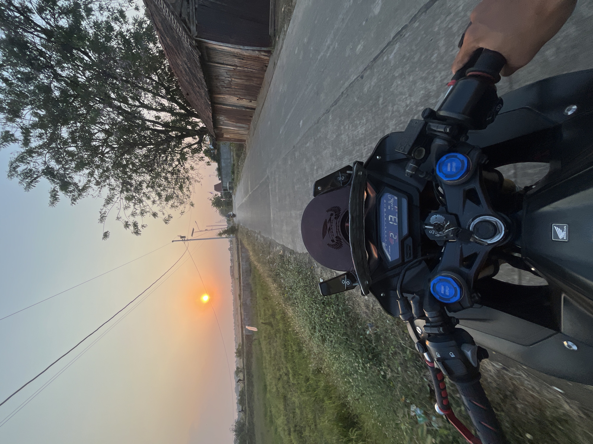 This is a first-person perspective, eye-level, medium shot of a motorcycle rider on a paved road in Pecangaan, Indonesia, at sunset. The rider's hands are visible on the handlebars. The motorcycle's digital dashboard displays "42" and "2 13". To the left of the road is a green, grassy field with some vegetation. To the right of the road, there is a dilapidated building with a corrugated metal roof and siding. In the background, the sun is low in the sky, casting a warm glow. A large tree is also visible to the right of the road. Overhead power lines stretch across the frame.
