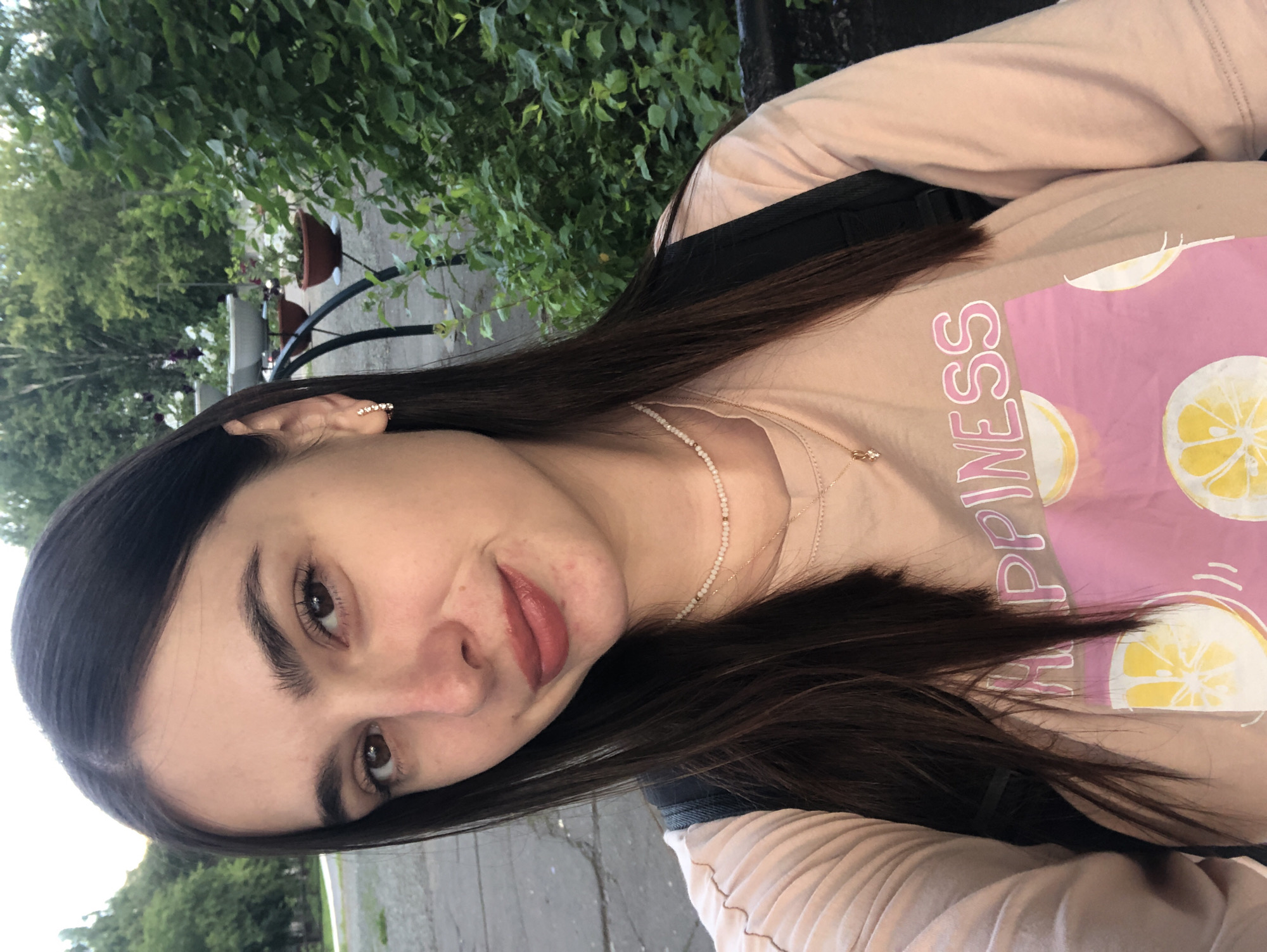 A young woman with dark brown hair and brown eyes smiles at the camera in an outdoor setting. She is wearing a light peach-colored long-sleeved shirt with a pink graphic that includes the word "HAPPINESS" and illustrations of sliced lemons. She also wears a delicate pearl necklace and a small gold pendant. The background features lush green foliage, potted plants, and a paved walkway. The overall scene suggests a casual, daytime moment.
