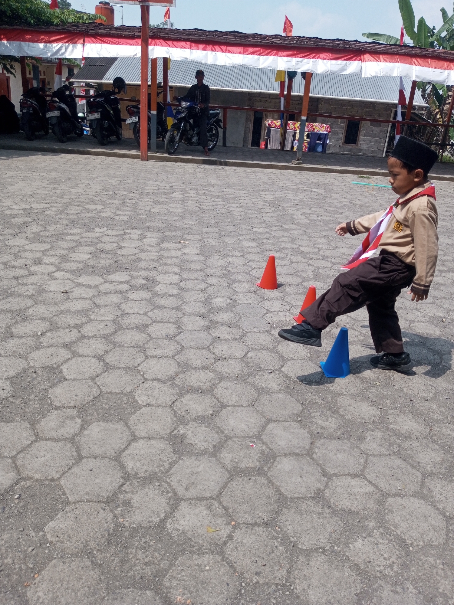 In the city of Kalibebe, Indonesia, a young boy dressed in a scout uniform, complete with a brown uniform, dark pants, and a black cap, is captured in motion. He is actively engaged in an outdoor activity, his right leg extended mid-kick, aiming towards a blue cone placed on the ground. The scene unfolds on a textured, hexagonal paved surface, indicative of a schoolyard or public plaza. Several other cones, two red and one blue, are strategically positioned around the boy, suggesting an obstacle course or a training exercise. In the background, a covered structure, likely part of a building, is adorned with red and white banners. Several motorcycles are parked nearby, and a man in dark clothing stands beside one of them, observing the activity. The overall atmosphere appears to be daytime, with a clear, bright sky overhead.