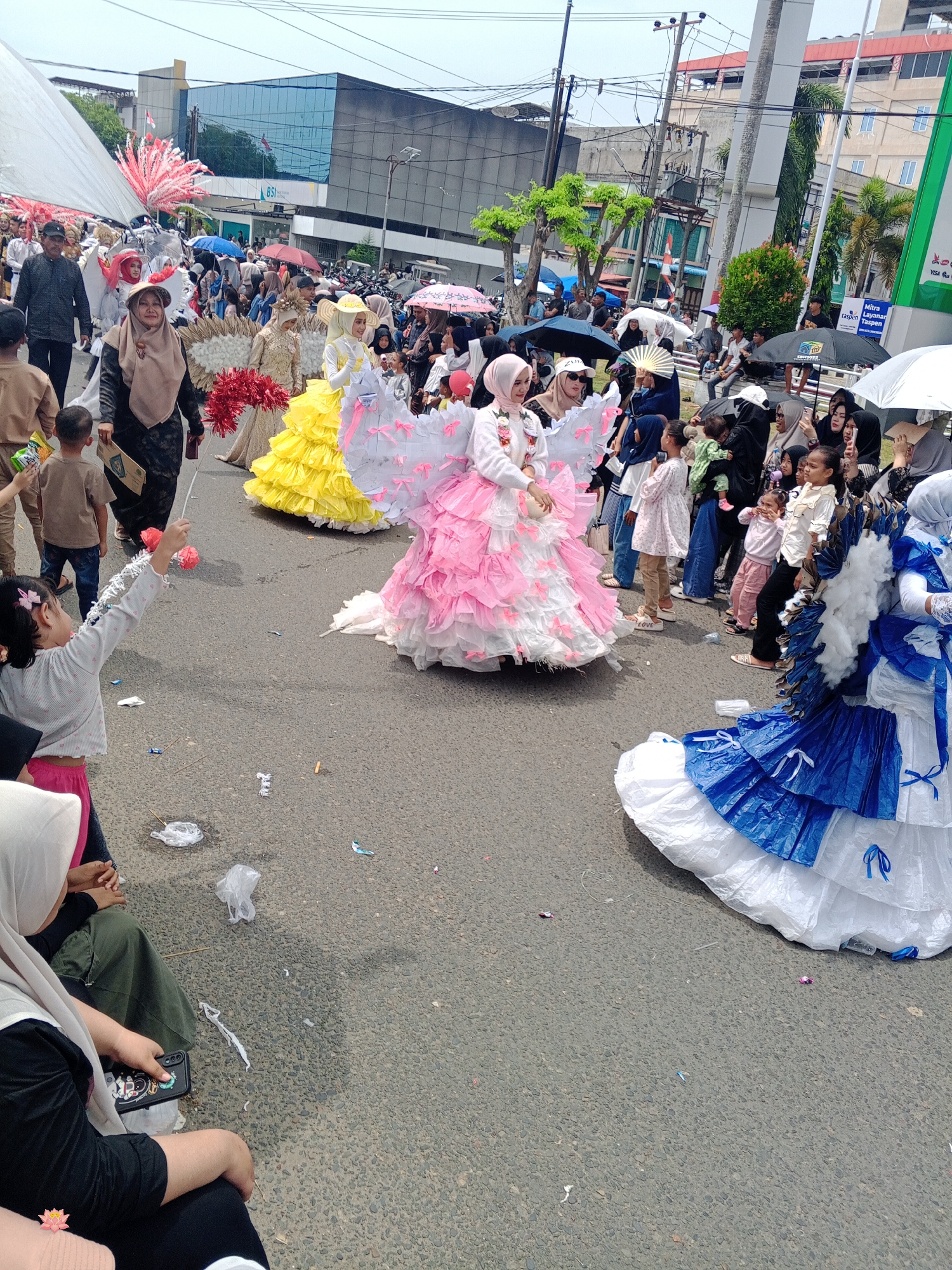 The image depicts a street scene in Lhokseumawe, Indonesia, likely during a parade or festival. A crowd of people, including children and adults, are present on a paved road. Several individuals are dressed in elaborate costumes, some resembling angels with large, feathery wings, and others in flowing, colorful dresses. One woman in a brown hijab and a black dress is holding a bouquet of red and white flowers. In the foreground, a young girl in a light-colored top and pink pants is holding a decorative wand. The background shows buildings, trees, and overhead wires, suggesting an urban environment. The ground is littered with some debris, including crumpled paper and plastic. Several people are holding umbrellas, possibly for shade or protection from light rain. The overall atmosphere appears festive and participatory.