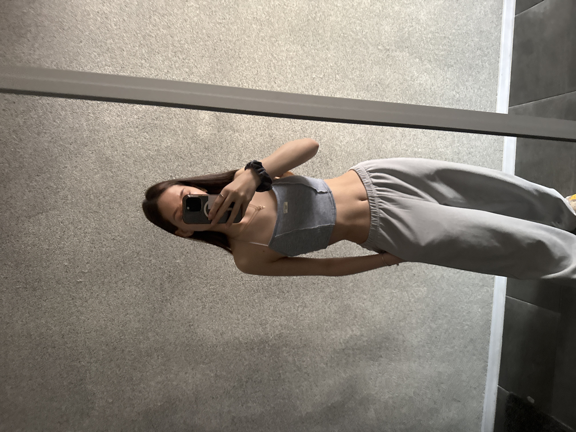 A person stands in front of a mirror, taking a selfie with a phone. The person is wearing a gray crop top and gray sweatpants. The background consists of a gray wall. The exact location within Sofia, Bulgaria, cannot be determined from the image.