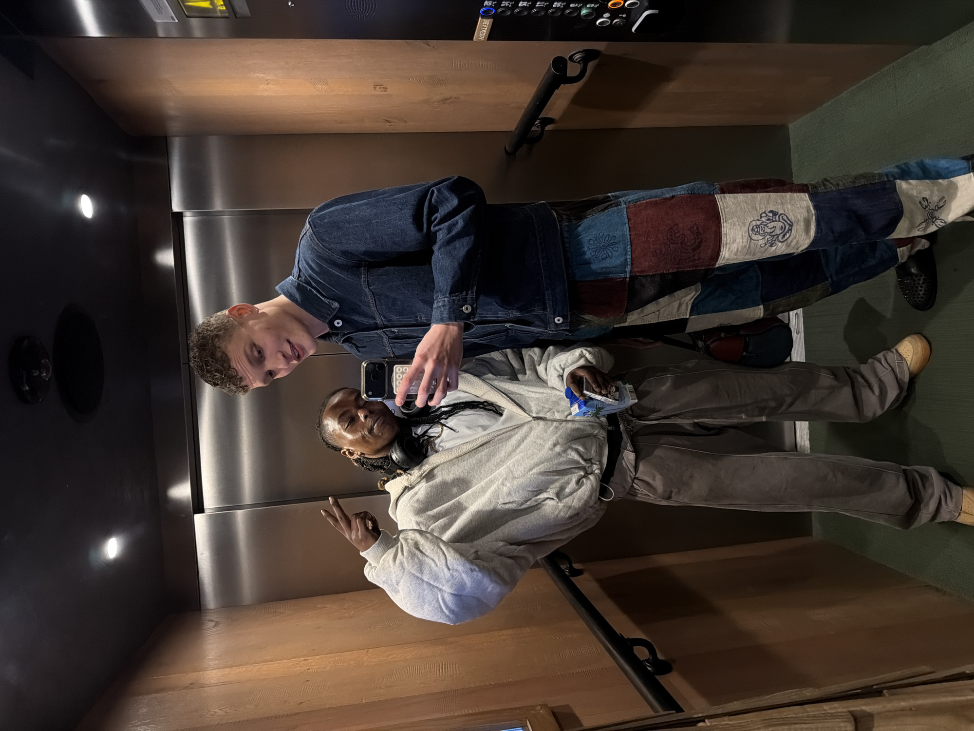 Two individuals are captured in a mirror selfie taken inside what appears to be an elevator, which features wooden paneling on the side walls, a dark metallic back wall with a reflective surface, and a greenish floor or lower wall section. The image is oriented vertically with subjects standing upright.
One person, with light skin and curly hair, stands on the right, wearing a denim jacket and distinctive patchwork pants adorned with various patterned fabric squares, some featuring embroidered designs such as a cartoon-like character. They are looking towards the camera (held by the other person) with a slight smile. Their right hand is partially visible, holding a smartphone.
The second person, with dark skin and braids, is on the left, holding a smartphone with both hands to take the mirror selfie. They are wearing a light grey zip-up hoodie over a white t-shirt, grey pants, and have headphones draped around their neck. This individual is making a peace sign with their left hand and looking towards the phone. A blue drink carton or bottle is visible in their right hand. A small bag is seen on the floor between the two individuals.
Notable features of the elevator include a black handrail mounted on the wooden wall and an elevator control panel with various buttons and indicators (e.g., '0', '00', 'RS') and what appears to be "LIFT TEST" written on a small plate beneath the buttons. The lighting suggests an indoor setting with artificial light sources. The overall context suggests the photo was taken in London, United Kingdom.