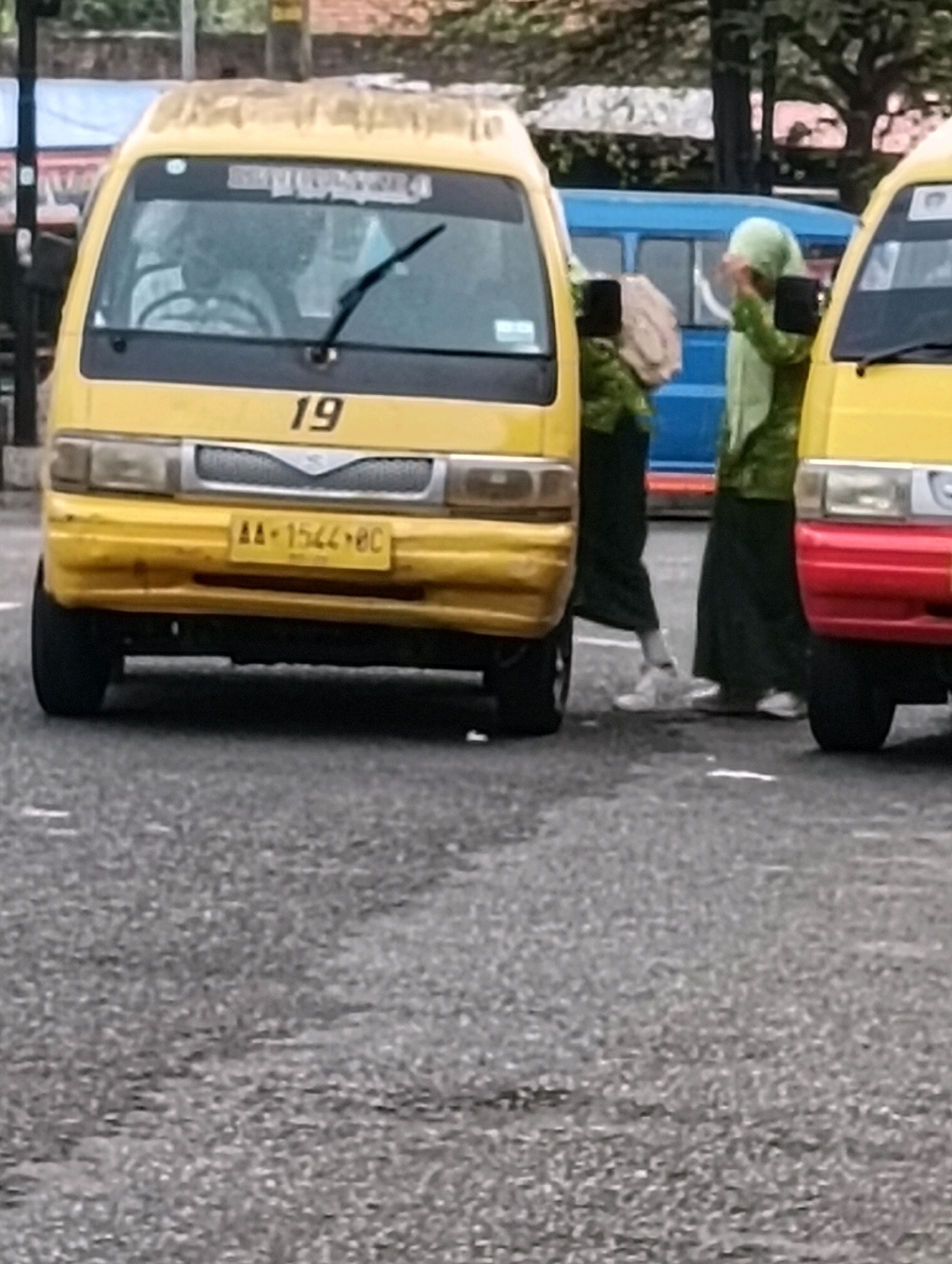 The image depicts a street scene in Purworejo, Indonesia, with two yellow vans and two individuals dressed in green clothing. The foreground features the front of a yellow van with the number "19" visible on its hood and a license plate that reads "AA-1544-BC". The van has its headlights on. To the right of this van, another yellow van is partially visible, showing its red bumper. Two individuals are standing between the vans. One, on the left, is wearing a green headscarf and green clothing, holding a light-colored bag. The other, on the right, is also wearing green attire and a headscarf, appearing to adjust it. In the background, a blue vehicle and trees are visible, suggesting an outdoor, possibly urban or semi-urban, setting. The ground is paved with asphalt. The overall impression is a snapshot of daily life in a street environment.