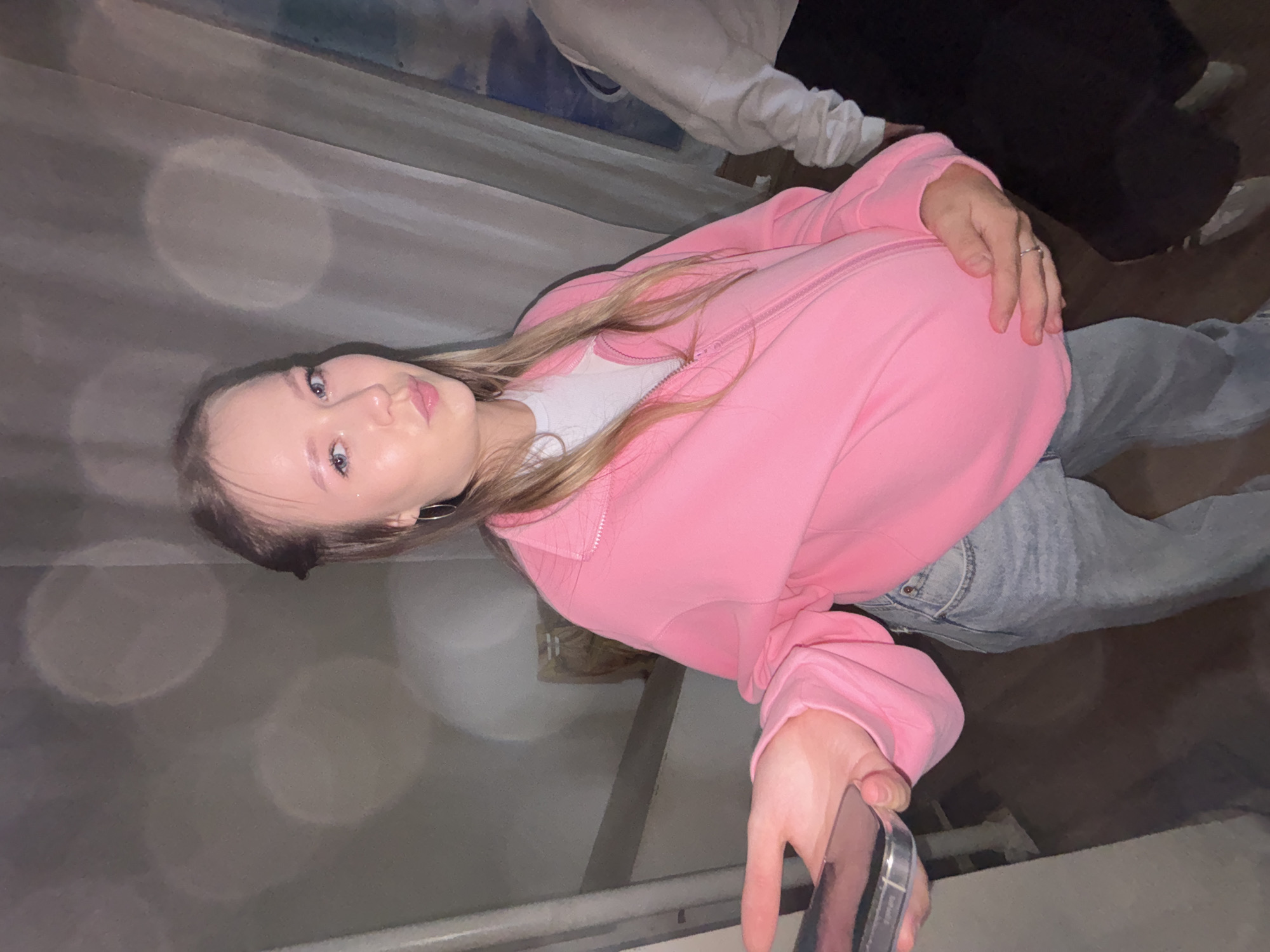 In Chaikovsky, Russia, a young woman with blonde hair, wearing a pink zip-up jacket over a white t-shirt and light blue jeans, poses for a photo indoors. She has on hoop earrings and a ring on her left hand, which rests on her abdomen. Her right hand is extended, holding a smartphone. The background shows out-of-focus circular lights, a wall, and curtains. Another person is partially visible to the right, wearing a light-colored top and dark pants. The overall lighting is dim.
