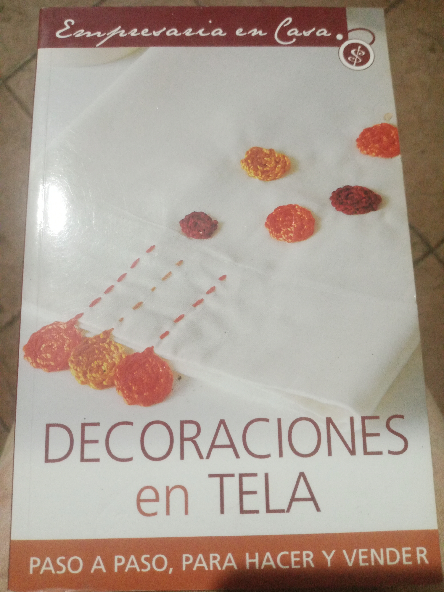 The image is a close-up of a book cover titled "Decoraciones en Tela" (Decorations on Fabric). The cover features a white fabric background with several small, crocheted or embroidered embellishments in shades of red and orange scattered across it. The title is printed in large, maroon sans-serif font at the bottom of the cover, with "en TELA" in a slightly smaller font below "DECORACIONES". Above the main title, in a smaller, stylized script, the words "Empresaria en Casa." (Businesswoman at Home) are visible, accompanied by a small circular emblem containing a dollar sign. The bottom portion of the cover has a solid orange band with the text "PASO A PASO, PARA HACER Y VENDER" (Step by Step, To Make and Sell) in white capital letters. The background of the image shows the corner of what appears to be a table with a tiled surface. There are no people or animals visible in the image. There is no information in the image that indicates a location in the city of Santa Ana, Venezuela.