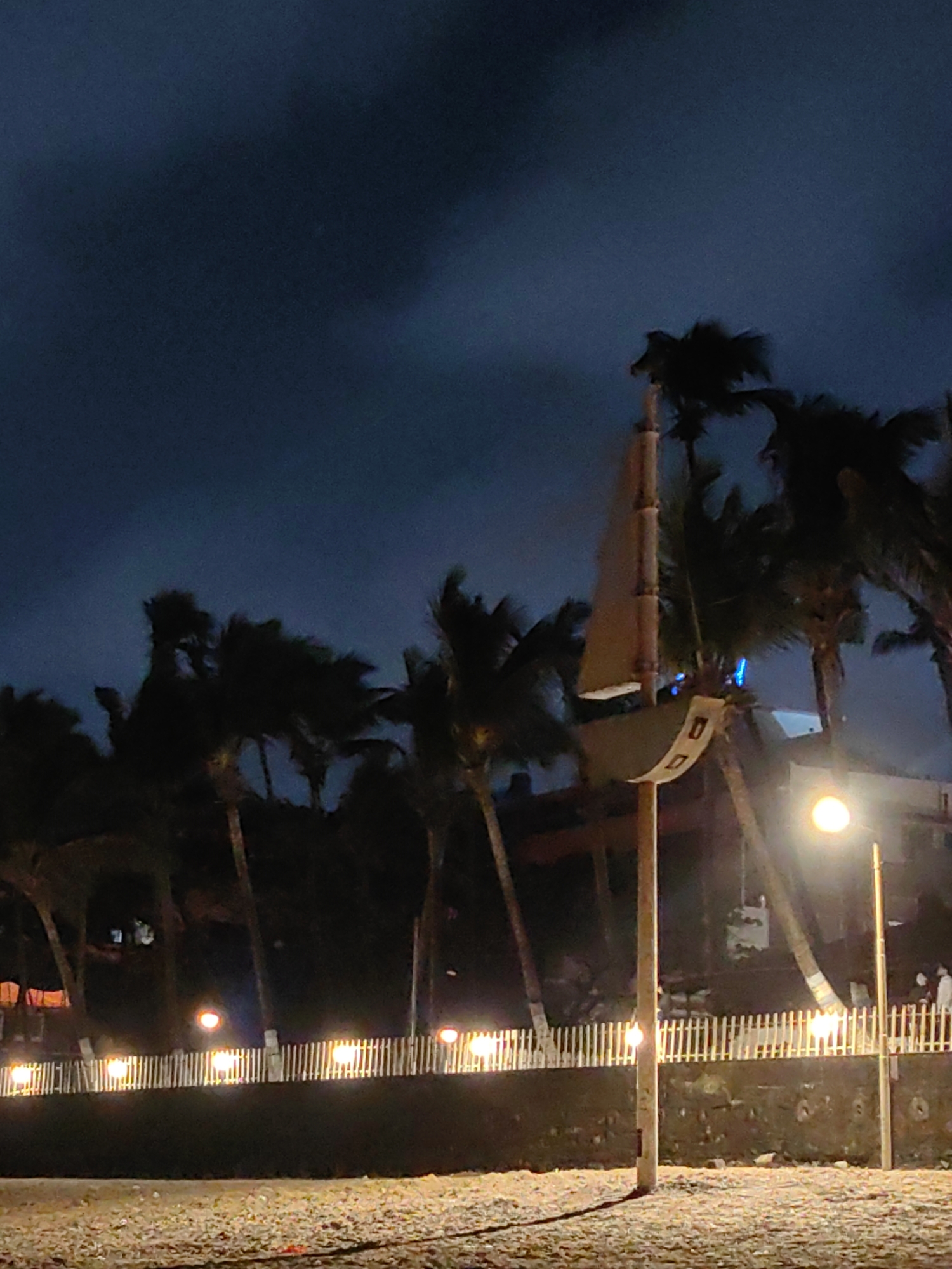The image captures a nighttime scene at Girgaum Chowpatty Beach in Mumbai, India. The foreground is a sandy beach, with some scattered debris. Beyond the sand, a low, dark wall separates the beach from a pathway lined with a white, picket-style fence. This fence is illuminated by a series of small, warm lights, creating a festive atmosphere.
Dominating the mid-ground is a tall, wooden pole topped with a decorative, boat-like structure, possibly a weather vane or a symbolic art installation. This structure has a sail-like element and a curved hull shape. Behind this, a row of silhouetted palm trees stretches across the scene, their fronds appearing windswept against the dark, cloudy sky.
In the background, various buildings are visible, some illuminated with lights, suggesting the presence of shops, restaurants, or other establishments along the beachside. A bright, circular street lamp stands to the right of the frame, casting a strong pool of light. Further in the background, a few indistinct figures can be seen near the illuminated buildings. The overall ambiance is one of a popular nighttime gathering spot, with the natural elements of the beach and palm trees contrasting with the man-made lights and structures. The sky is dark and overcast, suggesting it might be late evening or early morning, or perhaps indicative of approaching rain.