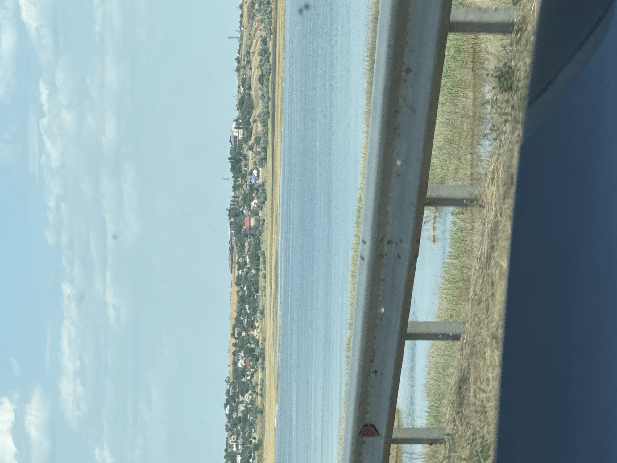 The image displays a body of water with a coastline dotted with houses and trees, likely a village. A metal guardrail runs along the foreground, separating the viewer from the water and the marshy vegetation by the shore. The sky is partly cloudy with a light blue hue. This scene is consistent with the coastal area of Koshary, Ukraine, which is known for its proximity to the Black Sea and its picturesque villages.