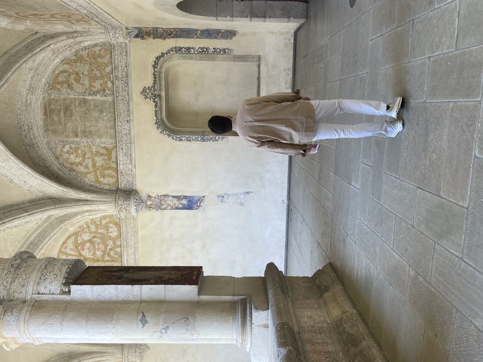 A man with dark hair, wearing a light-colored button-down shirt and white pants and shoes, stands in a colonnaded hallway. The hallway's walls are light-colored and adorned with decorative arches and painted ornamentation. The floor is tiled with large, light-colored stone slabs. The architectural style and ornamentation suggest a historical or religious building, possibly a monastery or church. The location is Oaxaca City, Mexico.