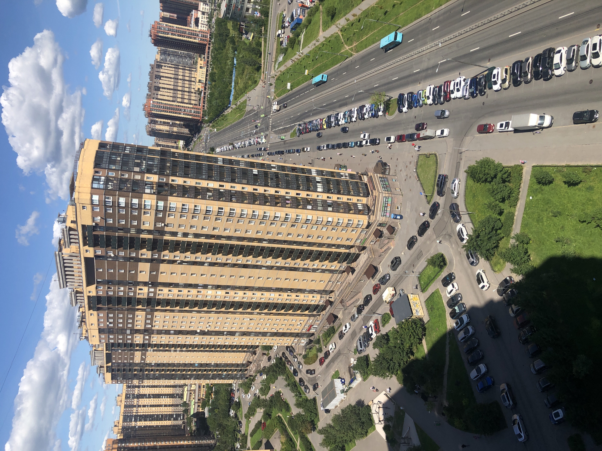 An aerial, wide shot captures a modern urban landscape under a bright, partly cloudy sky. Dominating the foreground is a tall, tan-colored apartment building with many windows and balconies. To its left, similar, though slightly shorter, residential buildings are visible. A multi-lane road runs alongside these buildings, with numerous cars parked along its sides and driving on it. Two blue buses are seen on the road, one further down than the other. Green trees and grassy areas are interspersed throughout the scene, bordering the buildings and the road. In the distance, more residential buildings can be seen, creating a dense urban environment. The overall impression is one of a bustling, contemporary city with a focus on high-rise living and extensive roadways.