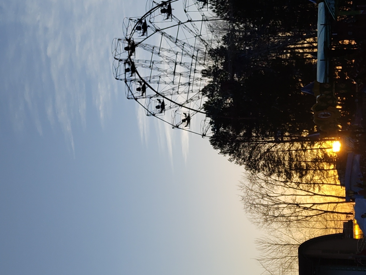The image shows a Ferris wheel against a backdrop of trees and a sky transitioning from blue to orange. Silhouetted figures can be seen in the Ferris wheel's gondolas. The sunlight filters through the trees on the right. The location is Chaikovsky, Russia.
