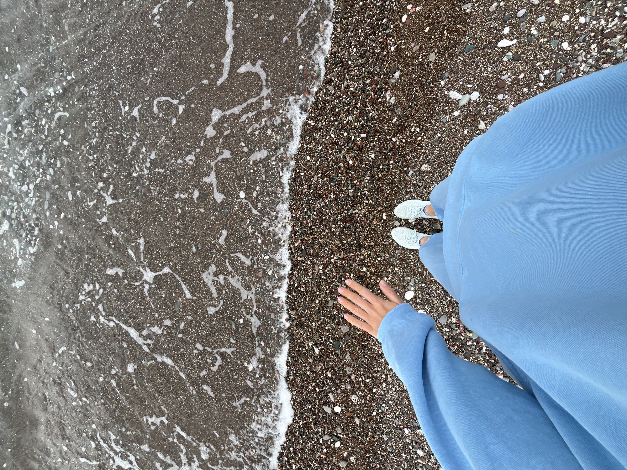 From an elevated, first-person perspective, the image captures a portion of a shoreline at Boreti, Montenegro. The left side is dominated by dark, turbulent water lapping onto the shore, with white foam tracing its path. To the right, the beach consists of small, dark pebbles interspersed with a few larger, lighter stones. A person stands on the pebbles, wearing light blue clothing that takes up a significant portion of the right side of the frame. The person's right hand is extended towards the shore, while a pair of white sneakers are visible on their feet.