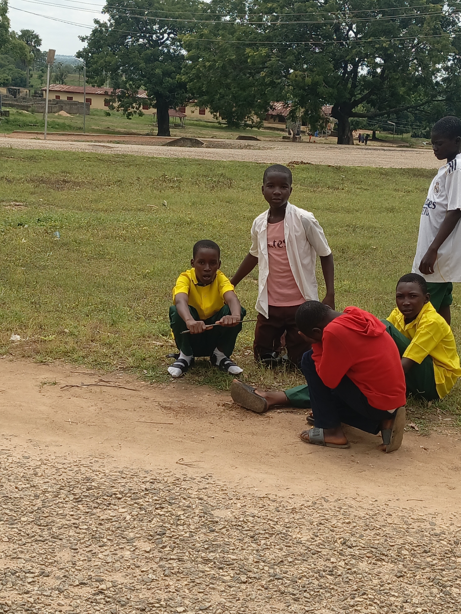 The image depicts a group of five young African children in an outdoor setting that appears to be a school or community ground in Akwanga, Nigeria. The children are dressed in casual clothing, with three of them wearing school uniforms. Two children are seated on the ground, one in a yellow shirt and green pants, holding a stick, and another in a red hoodie and dark pants, with his head down. Another child in a yellow shirt and green pants is crouched beside them. Two other children stand nearby, one in a white shirt and the other in a white and black jersey. The background shows buildings, trees, and a cloudy sky. The ground is a mix of dirt and gravel.