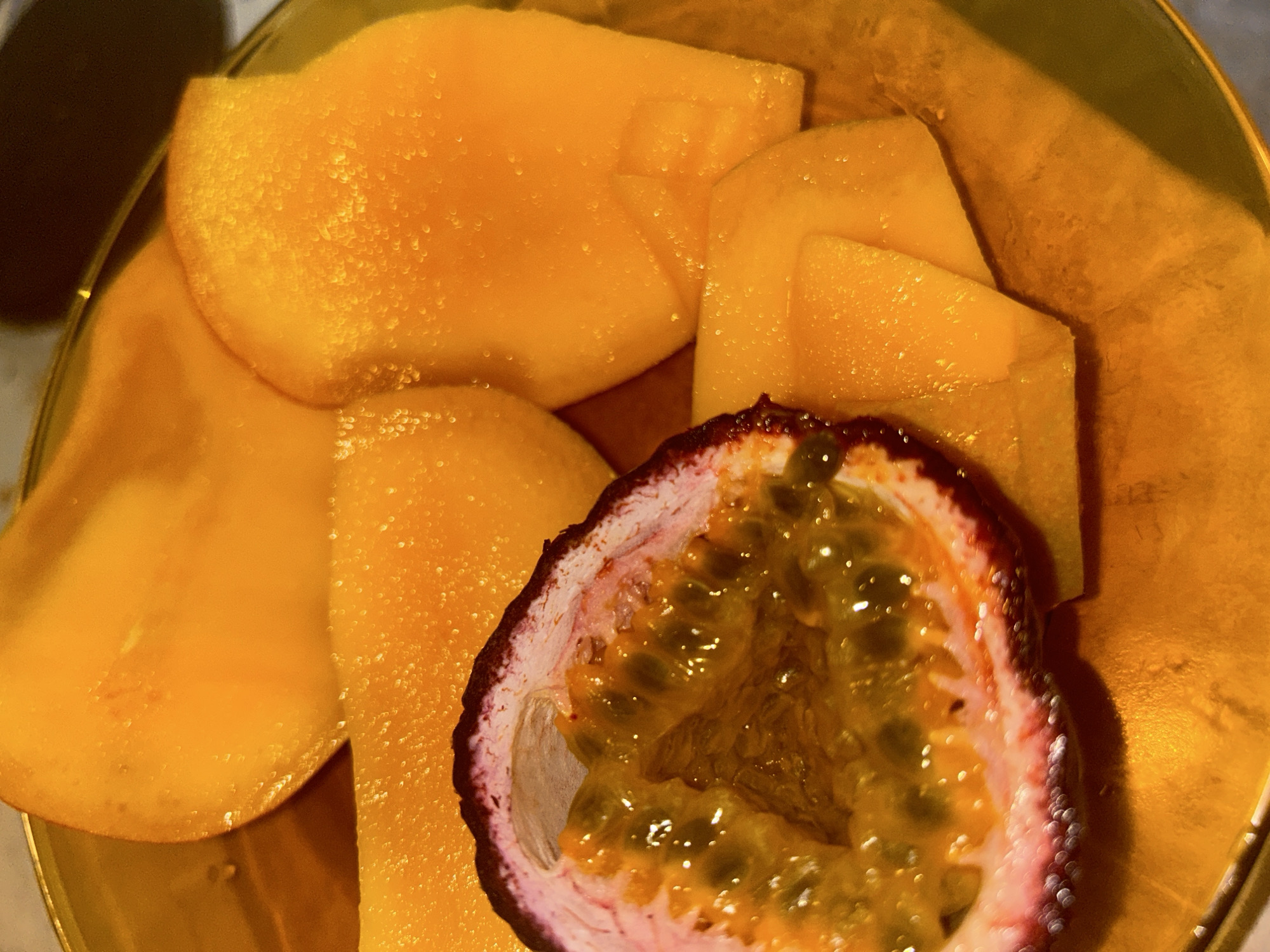 A close-up shot depicts a plate of sliced mango and a halved passion fruit. The mango pieces are arranged around the passion fruit, which is cut open to reveal its seeded pulp. The plate appears to be made of gold or a gold-colored material, and there is no indication of the specific location being Varna, Bulgaria. No people are visible in the image.