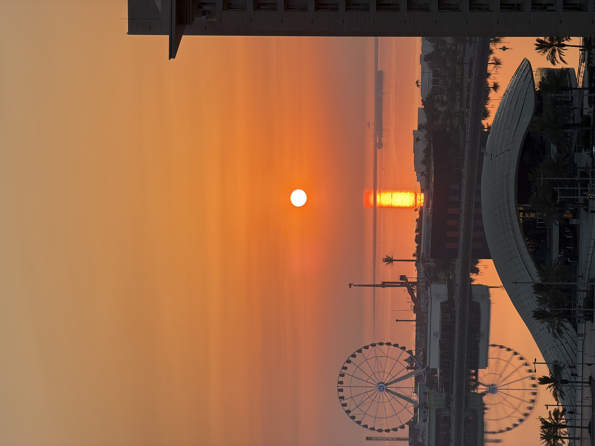 This is an exterior, eye-level, vertical, medium shot of a city waterfront at sunset. The sun is setting over the water, casting a bright orange glow across the sky and reflecting on the water. In the foreground, a large building with a curved roof and palm trees are visible. Behind it, there is a Ferris wheel and other structures along the waterfront. A large ship is visible on the horizon. The sky is a gradient of orange and yellow, with the sun being a bright white orb. The water is a deep orange, with a reflection of the sun and the ship. The buildings are silhouetted against the sunset. There are no people clearly visible in the image.