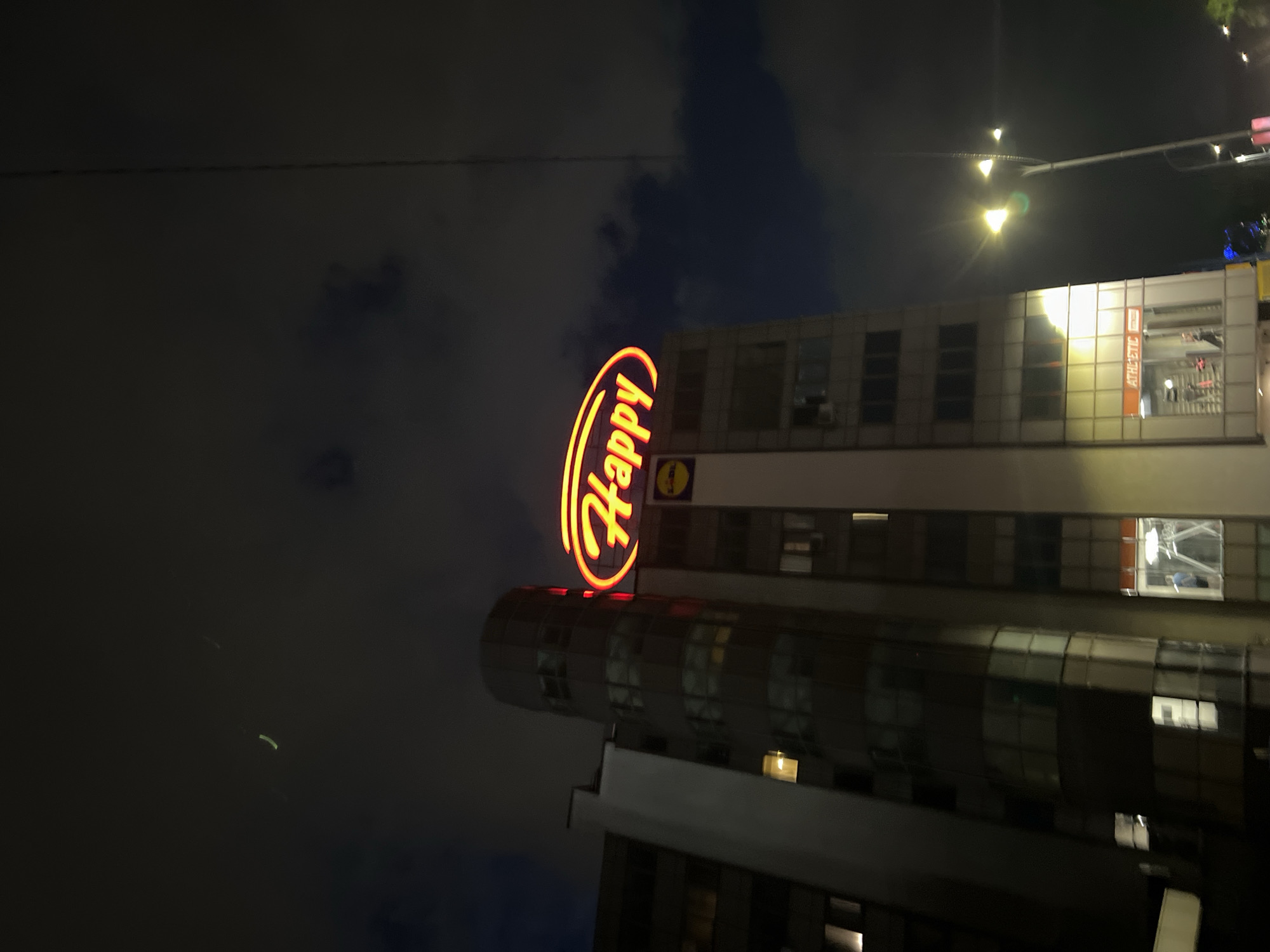 A nighttime view shows a tall building in Sofia, Bulgaria. The building has a large, curved neon sign reading "Happy" in yellow and red. The building appears to be a restaurant or entertainment venue, indicated by the sign and lights visible through the windows. A street light is visible to the right of the building. The sky is dark with some clouds.