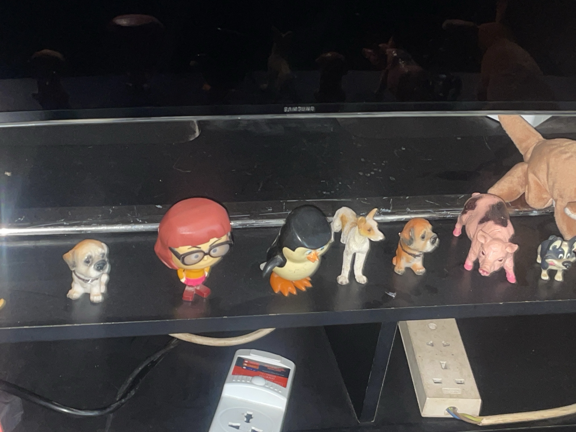 The image displays a shelf with several small figurines. From left to right, the figurines are a light brown dog, a female character with red hair and glasses, a black and yellow penguin, a white and tan dog, a brown dog, a pink pig, and a black and white dog. The shelf is black and appears to be made of particle board. Behind the shelf, there is a black reflective surface, possibly a television screen, showing some blurry reflections of objects that are not clearly identifiable. Below the shelf, there is a white electrical adapter with a power cord plugged into it. The overall lighting in the scene is dim. There are no people present in the image. The image is a photograph taken indoors. The city of Jos, Nigeria, is not depicted or referenced in the image.