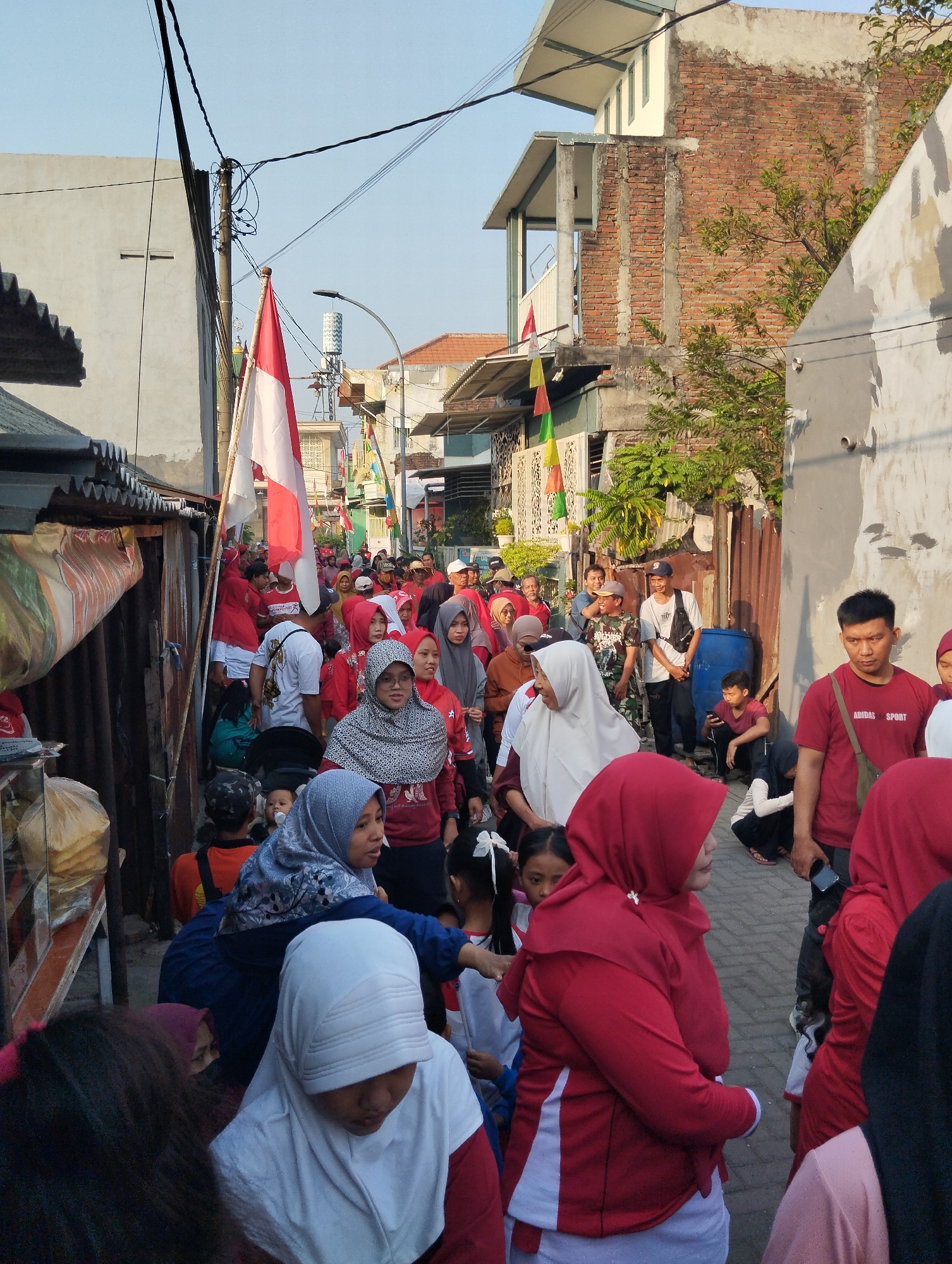 The image depicts a street scene in Berbek, Indonesia, likely during a festive or patriotic event, given the presence of Indonesian flags and numerous people wearing red and white attire. A procession is moving down a narrow street lined with buildings. In the foreground, a group of people are visible, including women wearing hijabs and children. Prominently displayed is a large Indonesian flag on a pole. The buildings appear to be residential, with some showing signs of age and construction material. The sky is clear, suggesting it is daytime. People are walking in the same direction, indicating a parade or march. Some individuals are holding their phones, possibly taking pictures or videos. The overall atmosphere appears to be one of community gathering and celebration.