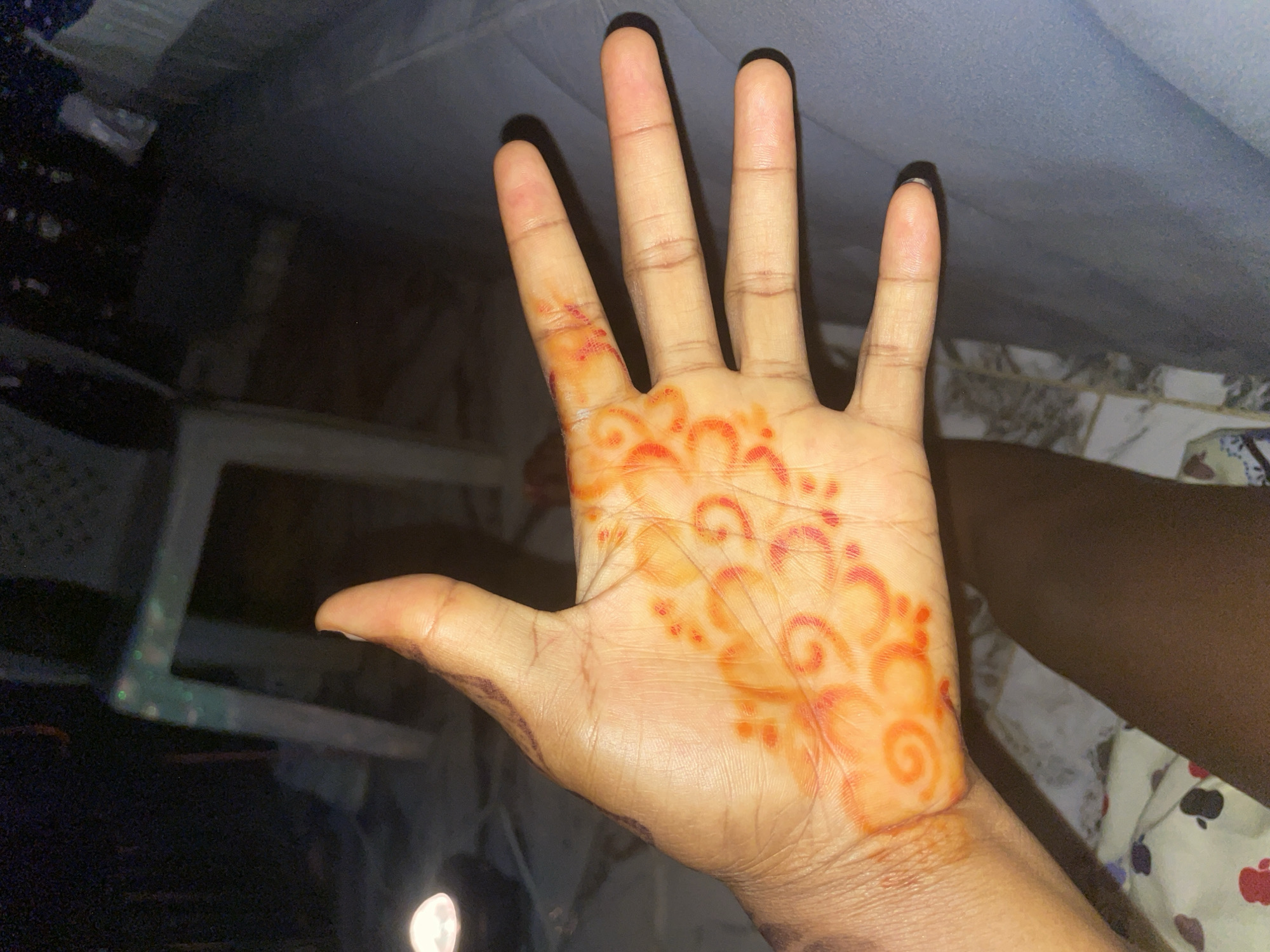 The image shows a person's hand with henna markings on the palm. The hand is light-skinned with a reddish-brown henna design featuring swirls and dots. The background is dark and out of focus, suggesting an indoor setting at night. A blurry, white object is visible in the upper left corner, possibly a lamp. A chair and part of a bed are also dimly visible. The overall impression is of a candid photograph taken in a dimly lit room. While the image itself does not contain explicit markers of Keffi, Nigeria, it is described as being taken there, suggesting a personal snapshot rather than a publicly documented scene.