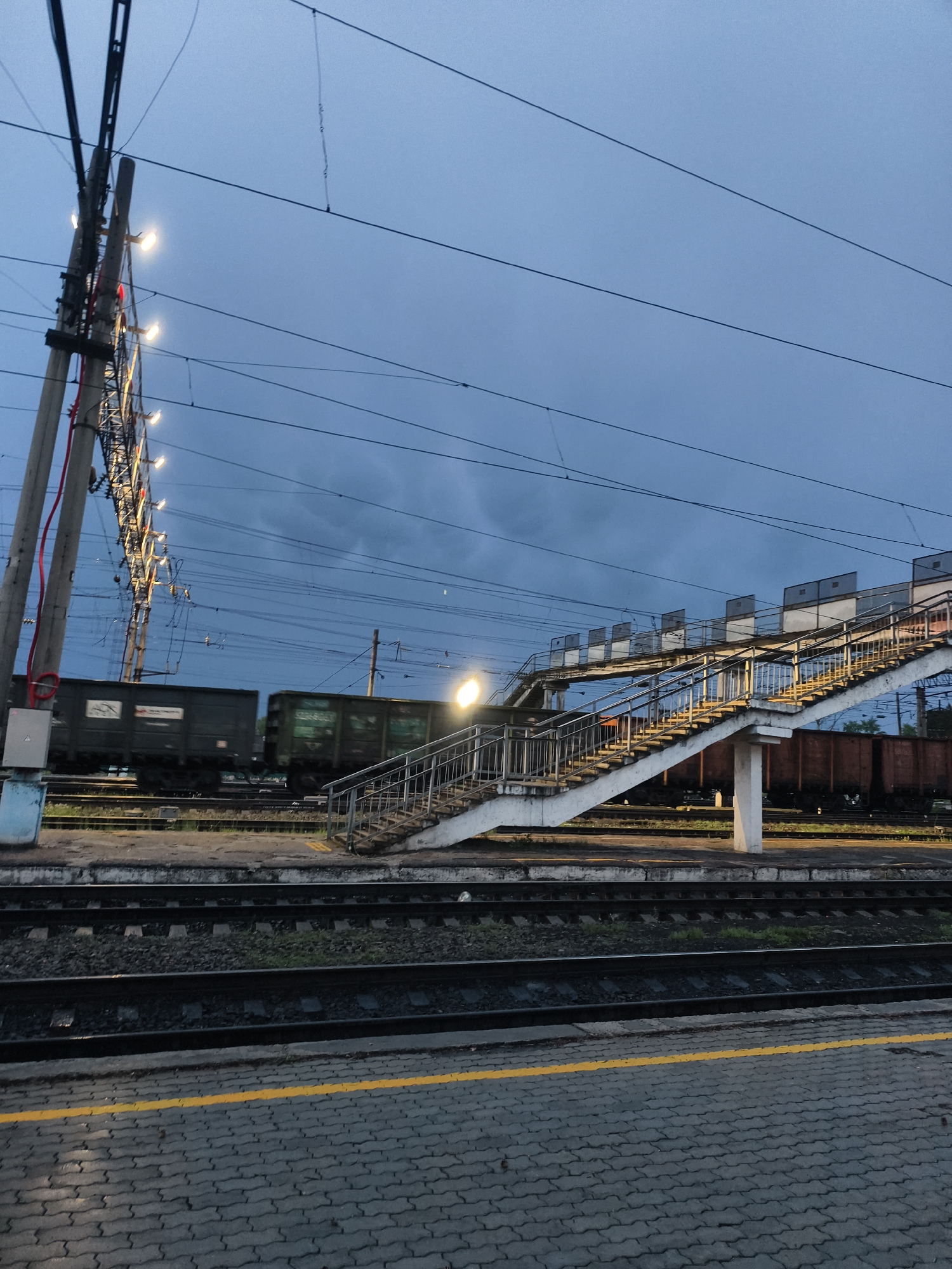The image depicts a train station in Magdagachi, Russia, at dusk. In the foreground, a platform paved with grey interlocking blocks features a yellow safety line. Train tracks run parallel to the platform. A pedestrian overpass with concrete stairs and metal railings spans the tracks. Several freight cars are visible on the tracks, some in motion, blurred by the camera. Overhead power lines crisscross the sky. A utility pole with illuminated lamps stands to the left. The sky is overcast and dark blue. No people are visible.