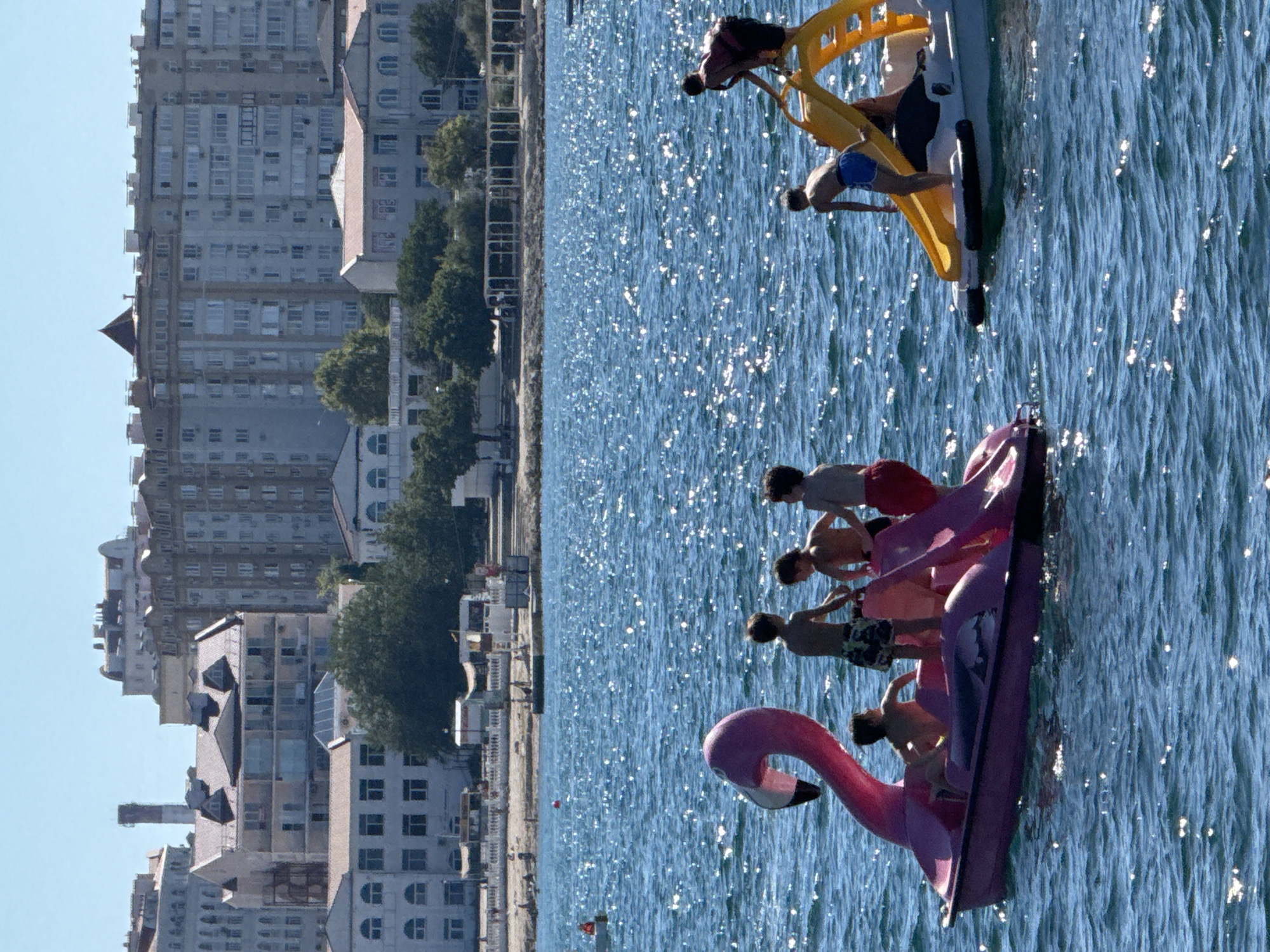 In Sevastopol, Ukraine, on a clear, sunny day, a body of water shimmers with reflected sunlight. In the foreground, a pink flamingo-shaped pedal boat is occupied by several young boys. One boy, wearing camouflage swim trunks, stands on the boat and appears to be handing something to another boy. Another boy, wearing red shorts, stands near the rear of the flamingo boat. A third boy, also in red shorts, is seated towards the front of the boat.
To the right of the flamingo boat, a white boat with a yellow slide is also carrying children. One boy, wearing blue swim trunks, is positioned on the slide's steps, preparing to go down. Another boy, wearing dark shorts, is climbing the ladder of the slide.
In the background, a shoreline is visible, with a promenade or walkway lined with trees. Beyond the trees, several multi-story buildings characteristic of a city skyline are present. The buildings vary in architectural style, with some appearing older and others more modern, featuring numerous windows and balconies. The sky above is a bright, cloudless blue.