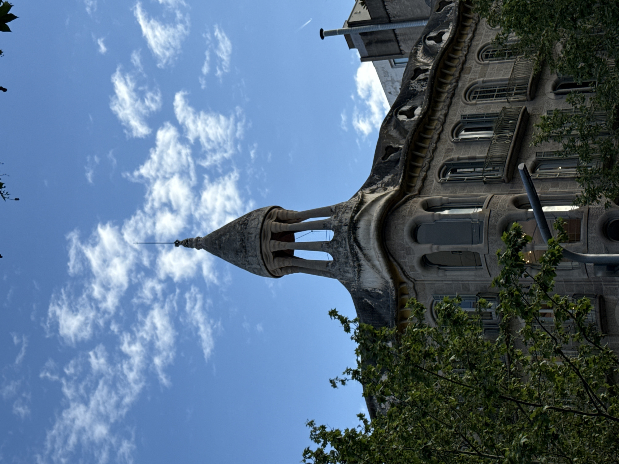 The photograph shows a section of Casa Milà, also known as La Pedrera, a modernist building designed by Antoni Gaudí in Barcelona, Spain. The structure is viewed from a low angle, emphasizing its organic, undulating facade and distinctive turret. The building's gray stone contrasts with the blue sky and scattered white clouds. A tree branch with green leaves is visible in the lower right corner of the image. The building has several windows and balconies. The distinctive architectural style is evident in the curved lines and unique details of the stone construction.
