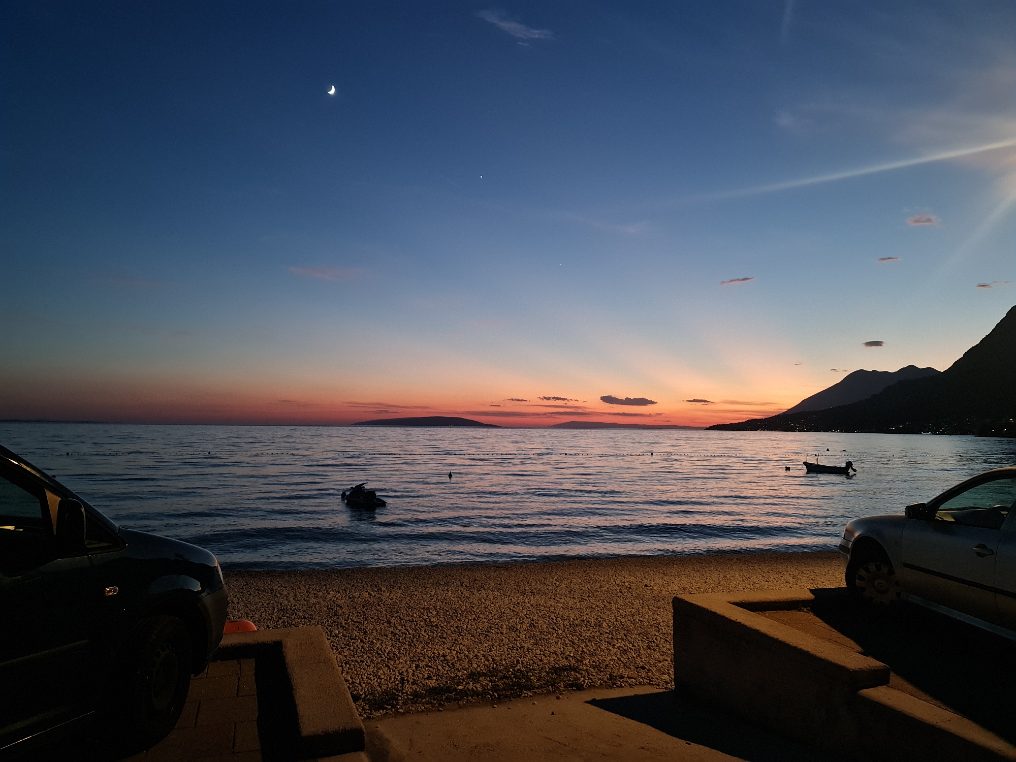 This image depicts a twilight scene at a beach in Općina Gradac, Croatia. The sky transitions from a deep blue at the top to shades of orange and pink near the horizon, where the sun has recently set. A crescent moon and two bright stars are visible in the upper left quadrant of the sky. Rays of light emanate from behind distant islands. The sea is calm with gentle ripples reflecting the colors of the sky. A jet ski is partially submerged in the water near the shore. In the foreground, a gravel beach is visible, with two cars parked on either side. The car on the left is a dark-colored SUV, and the car on the right is a silver sedan. A low concrete structure is present in the foreground, casting a shadow on the ground. The coastline is lined with dark mountains in silhouette against the illuminated sky.
