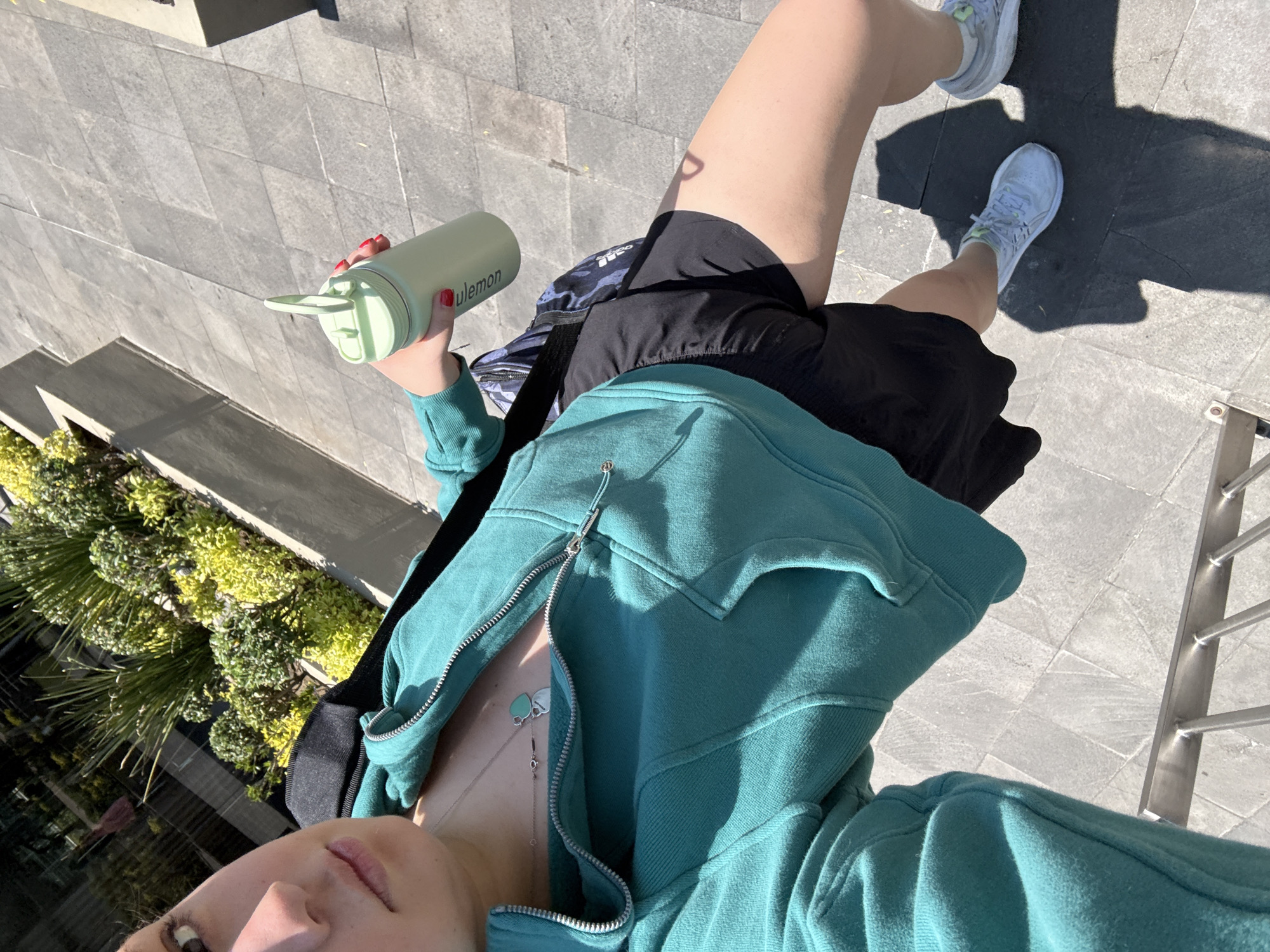 A high-angle, first-person perspective selfie captures a person standing outdoors on a bright, sunny day in Mexico City, Mexico. The individual, seen from the chest down, wears a teal or aqua-colored zipped hoodie, partially unzipped to reveal a silver necklace with two heart-shaped pendants (one teal, one silver). Black athletic shorts and light-colored athletic shoes are visible on their legs and feet.
The person's right hand, adorned with red nail polish, holds a light green "lululemon" branded water bottle with a straw lid. A dark backpack with a camouflage-like pattern and an "adidas" logo is slung over their shoulder, resting behind them.
The ground is paved with light grey square tiles, with some darker sections and distinct shadows cast by the person and surrounding structures, indicating strong sunlight. To the left, lush green and yellow-green foliage grows in a raised planter. A metallic railing is visible on the right side of the frame. The overall scene suggests an active, casual outdoor setting.