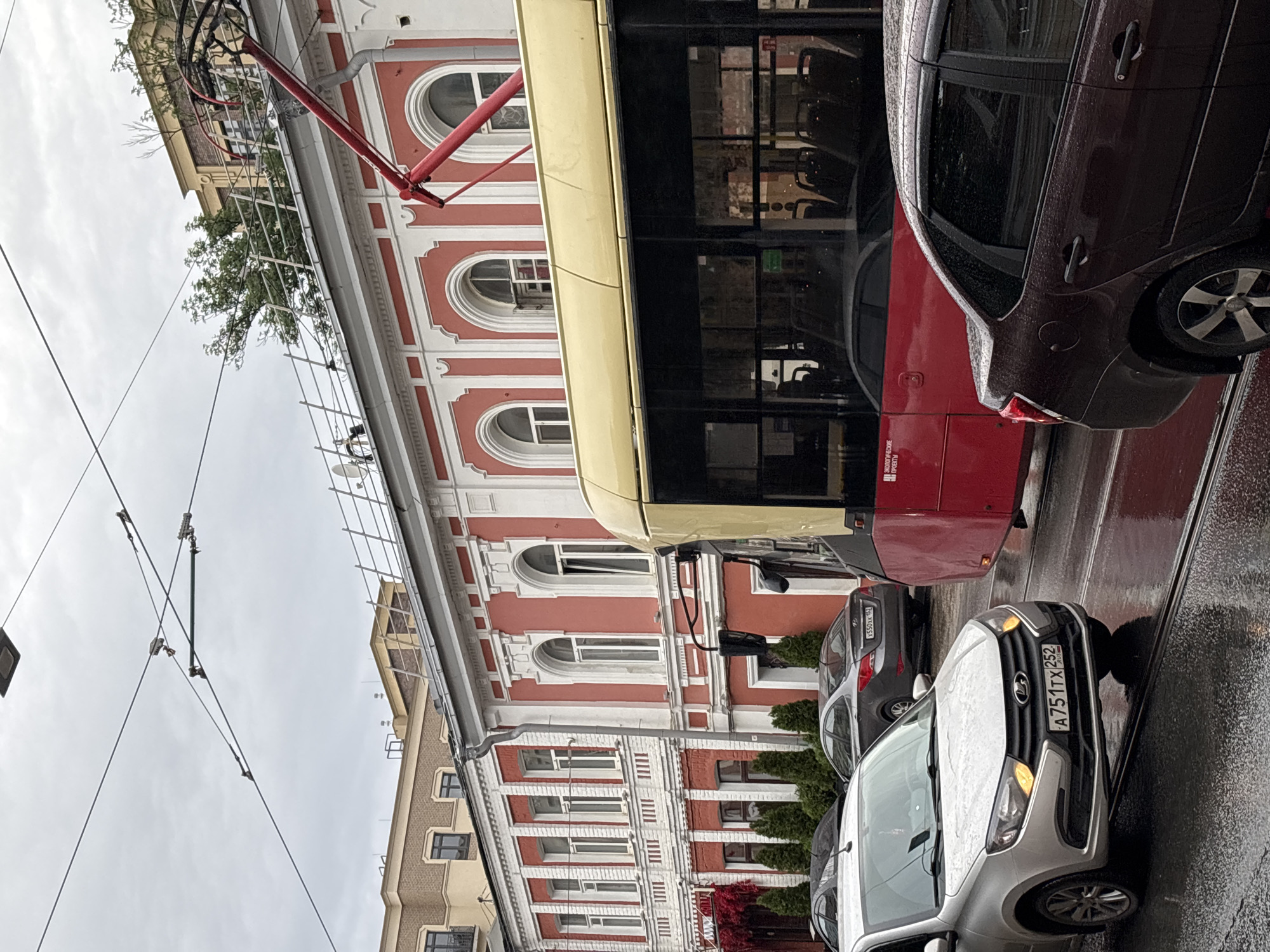 The image captures a street scene in Nizhny Novgorod, Russia, on a cloudy day. A red and yellow tram is the dominant subject, positioned on tram tracks in the foreground, obscuring some of the view of the street. To the left of the tram, a silver sedan with its headlights on is visible. Behind the silver car, a darker sedan is parked. The background features a red and white building with arched windows and decorative white trim. Overhead, power lines for the tram are strung across the sky. The street appears wet, suggesting recent rain. There are no visible people in the image.